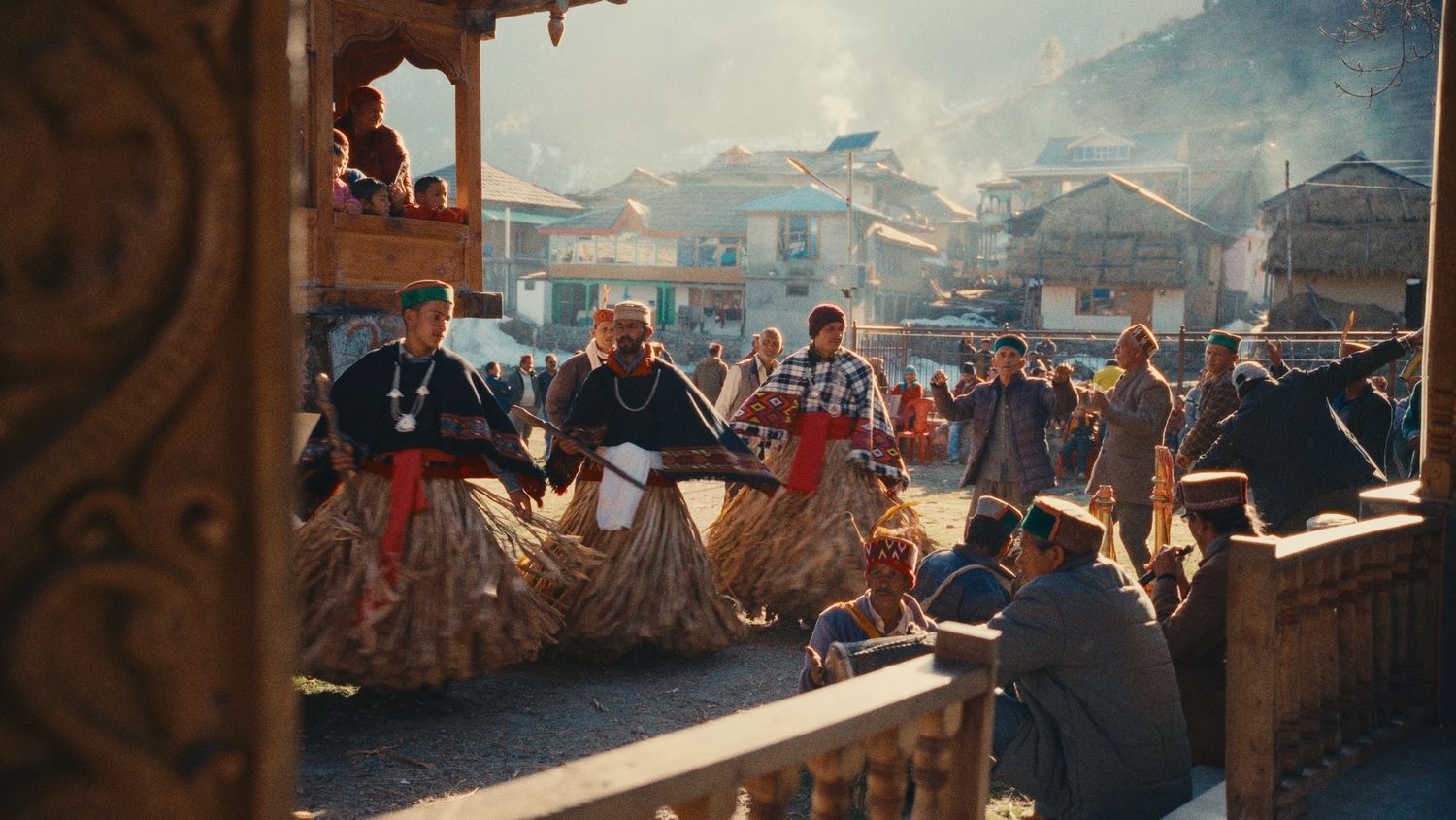 Traditional festival performers in colorful costumes dance in a sunlit mountain village square.