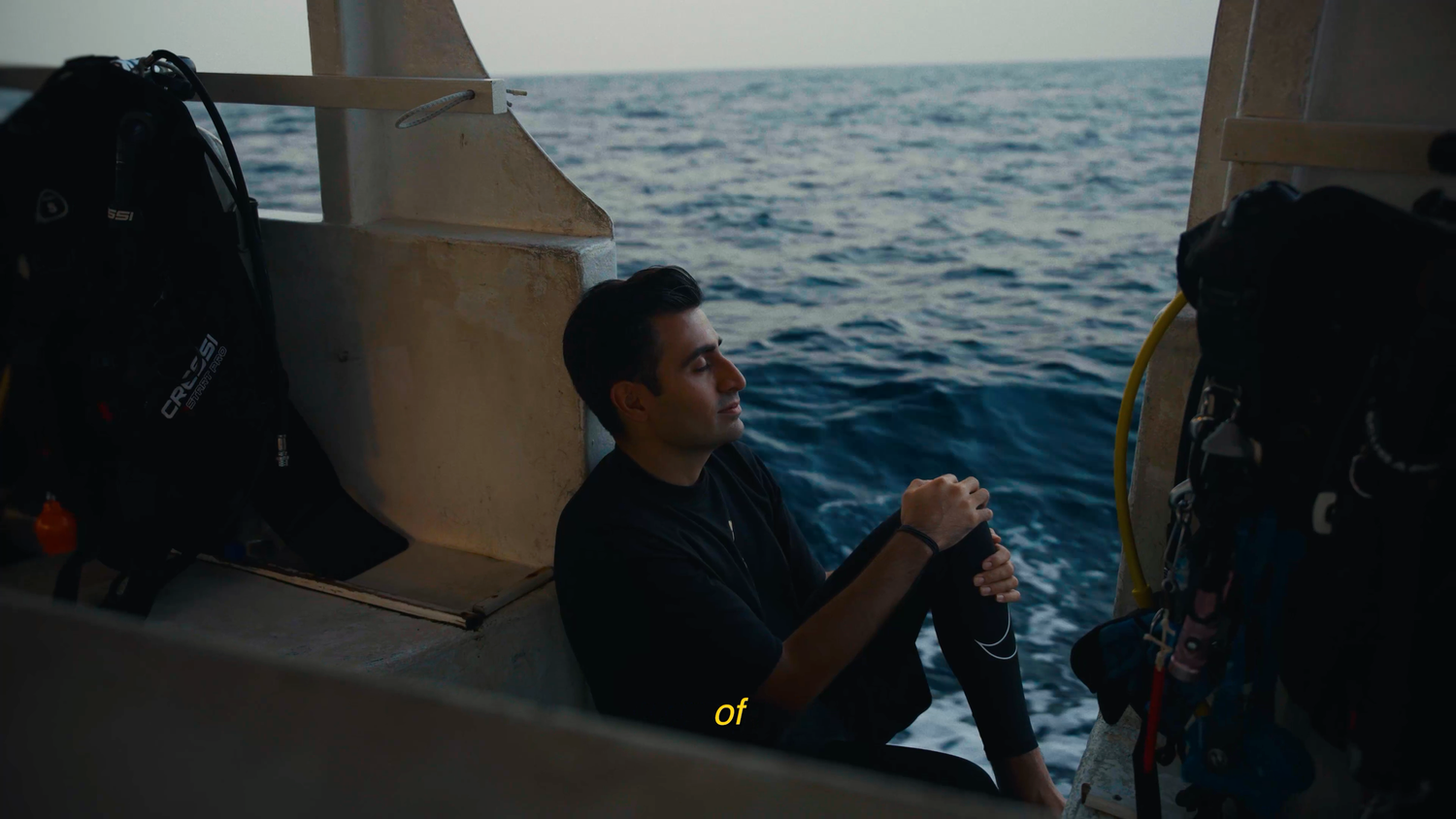 A man sits on a boat deck near scuba gear, smiling while holding equipment with the ocean in the background.