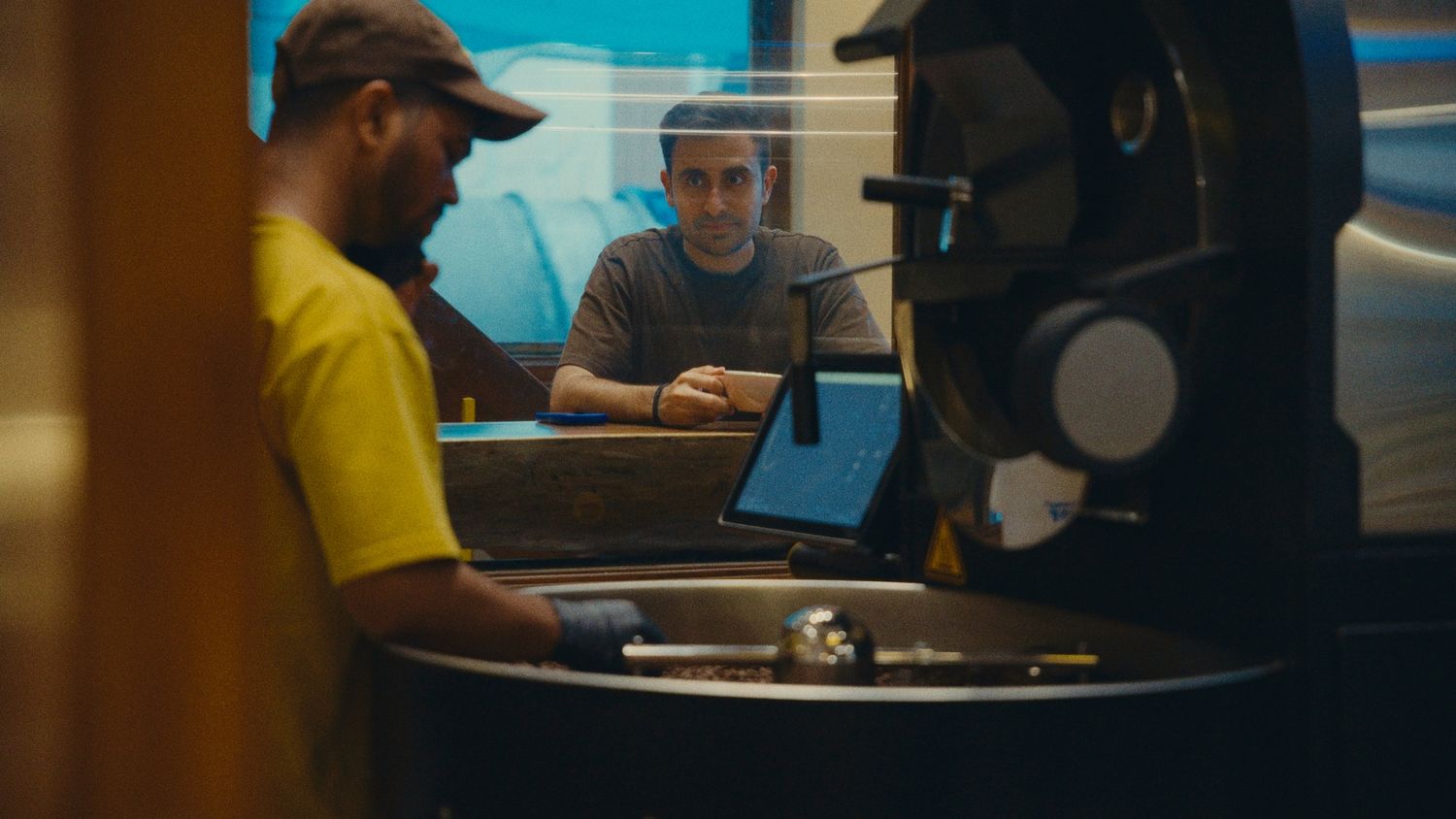 Two men working at a coffee roasting machine with a laptop in a warmly lit industrial cafe setting.