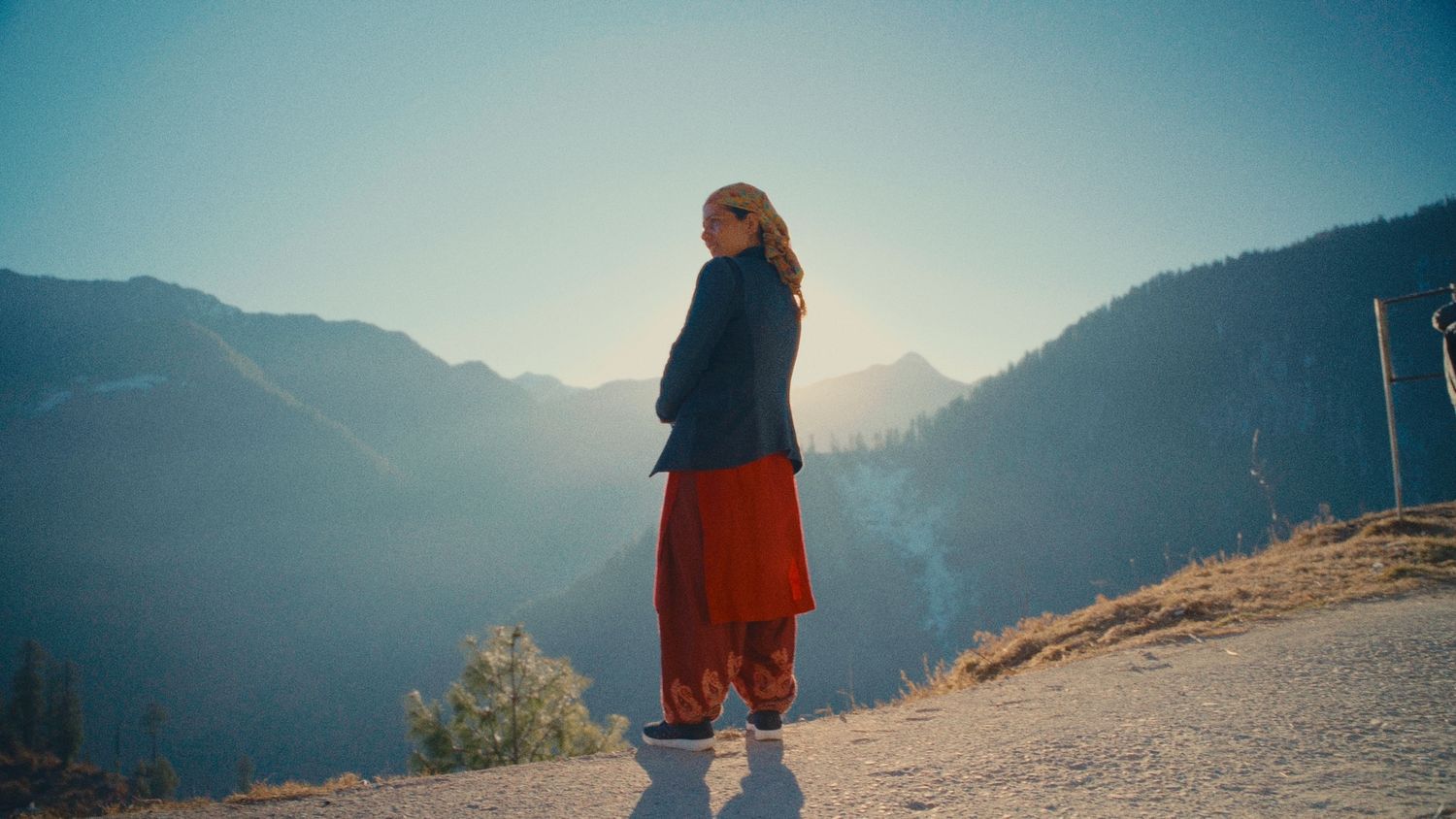 Woman in red pants and blue jacket stands on mountain overlook with scenic valley and sunlit peaks behind her.