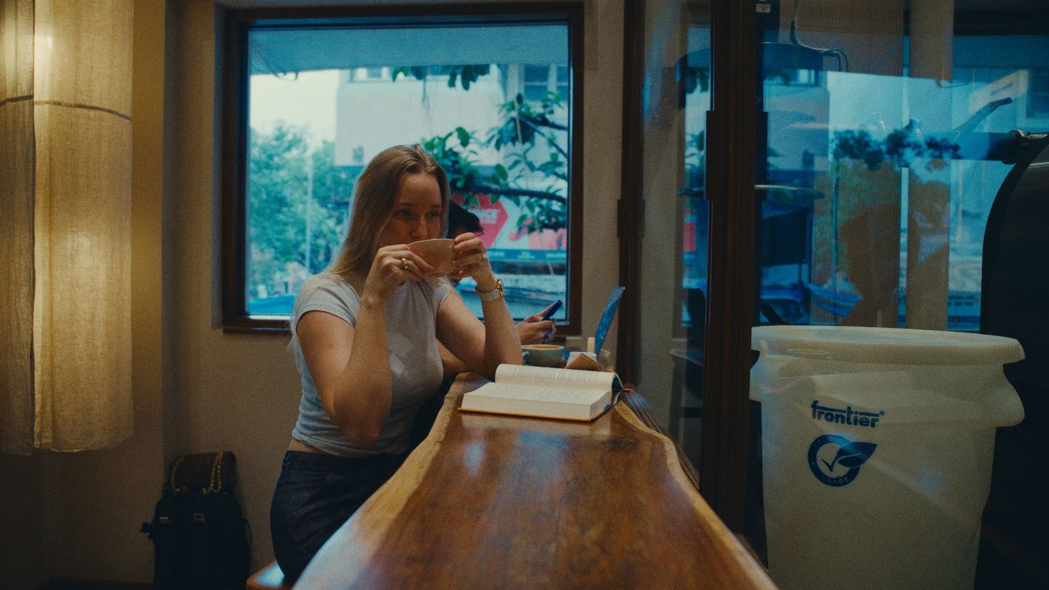A young woman sips a drink at a wooden bar counter near a window in a cozy cafe restaurant.