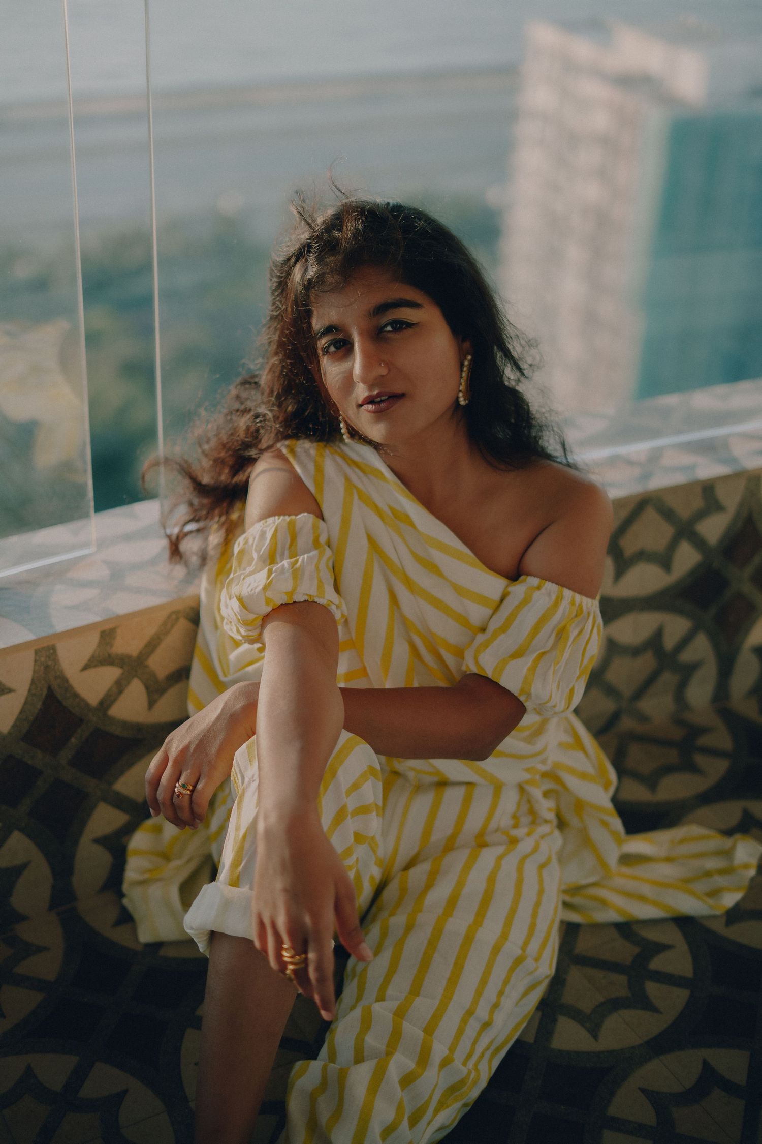 Model wearing yellow striped dress poses elegantly by window with natural lighting.
