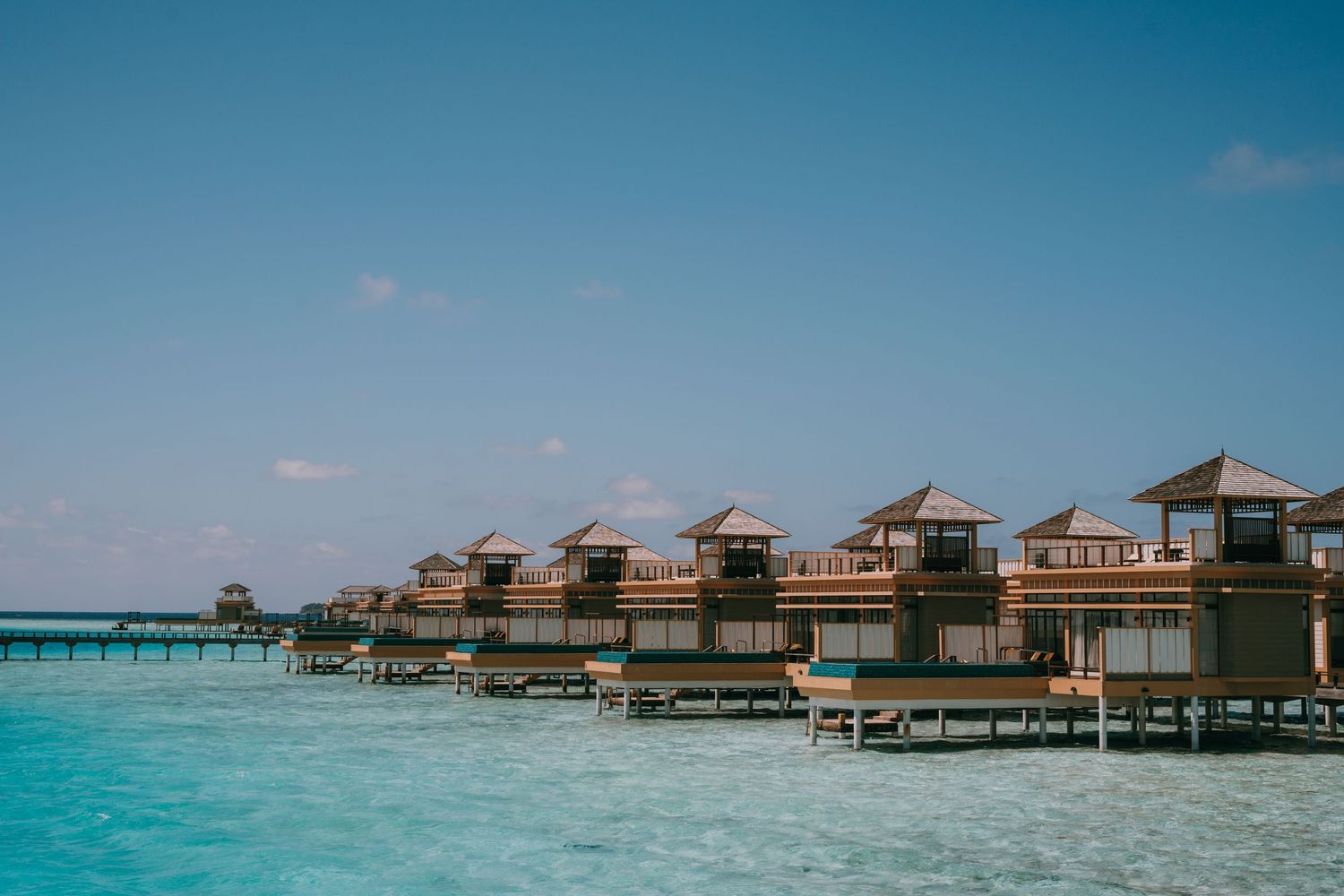 Row of overwater bungalows connected by wooden walkway over crystal clear tropical waters.