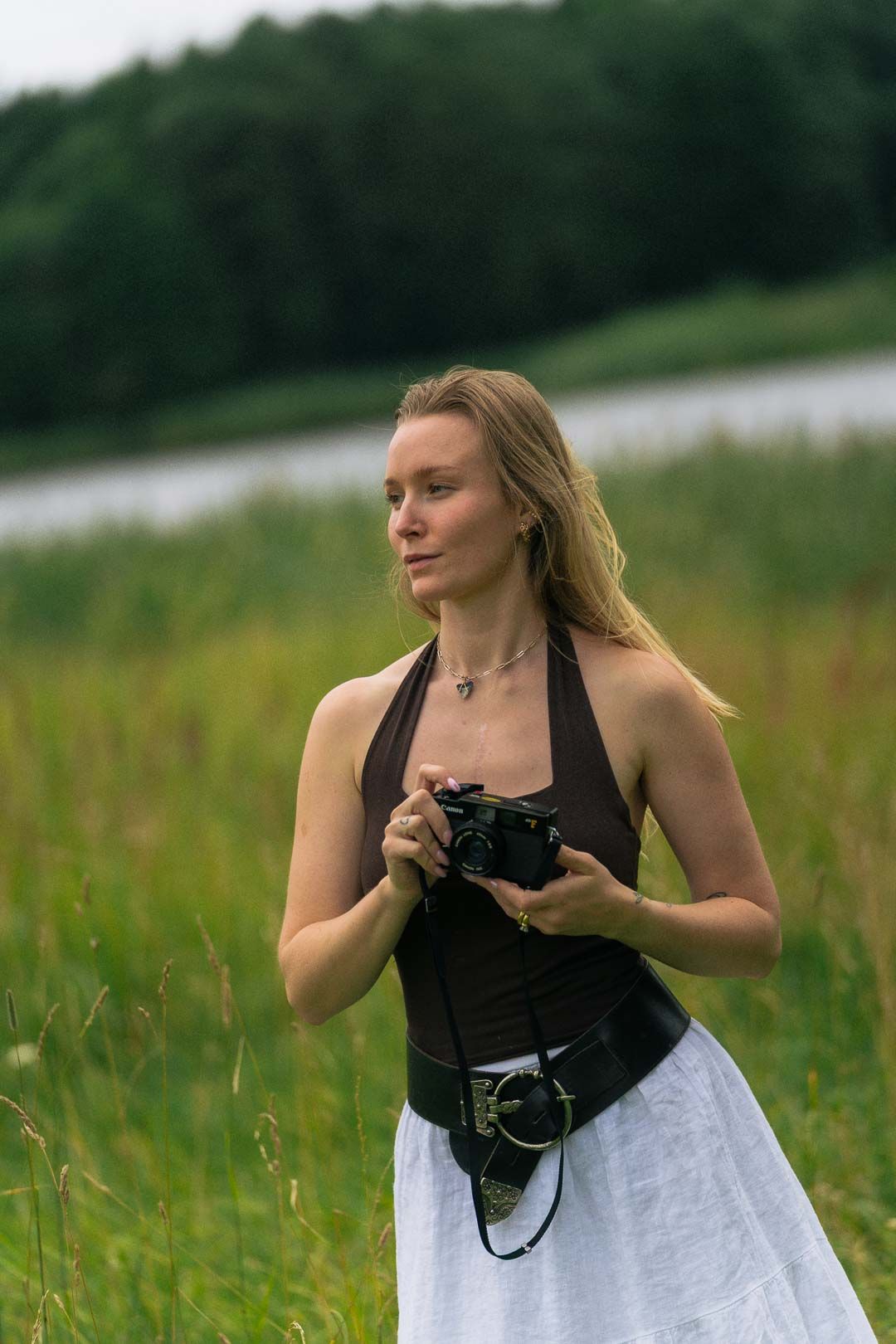 Young woman in brown halter top and white skirt holds vintage camera in lush green field near lake.