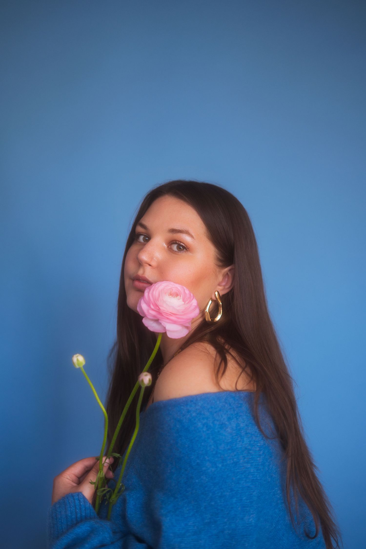 Portrait against blue backdrop with pink rose and gold hoop earrings.