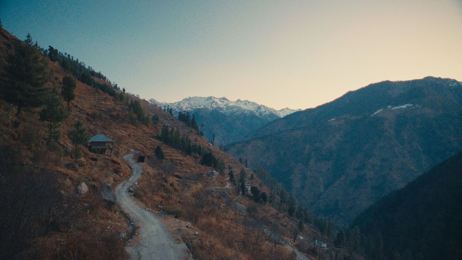 A winding mountain trail leads past a rustic cabin through autumn foliage toward snow-capped peaks at dusk.