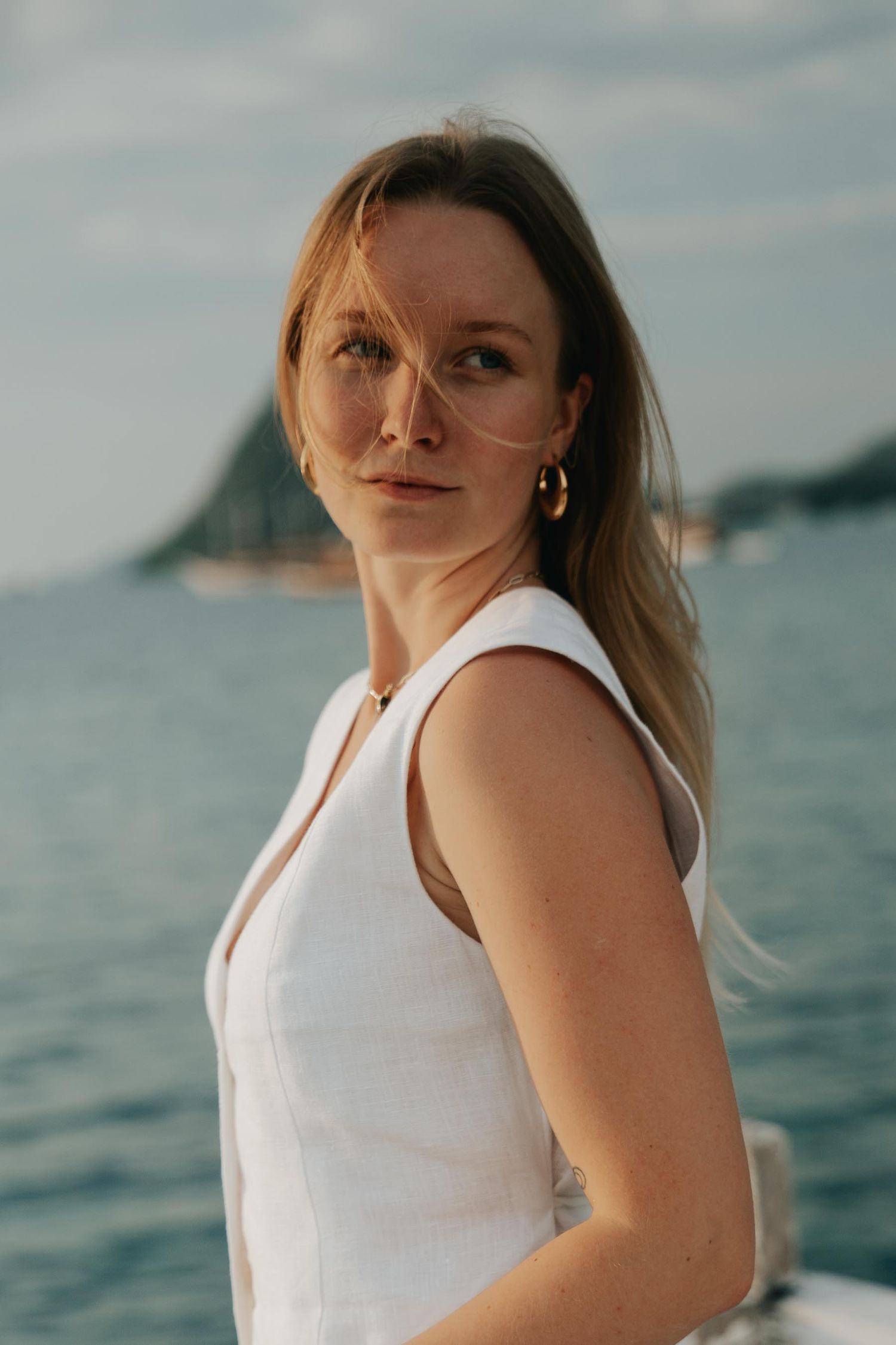 Seaside portrait series in white sleeveless top with ocean and mountains in background.