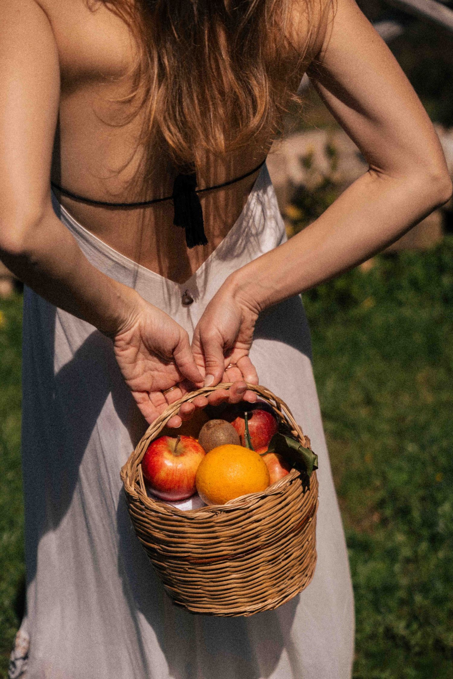 Woman in white dress holding a wicker basket filled with fresh apples and oranges outdoors in sunlight.