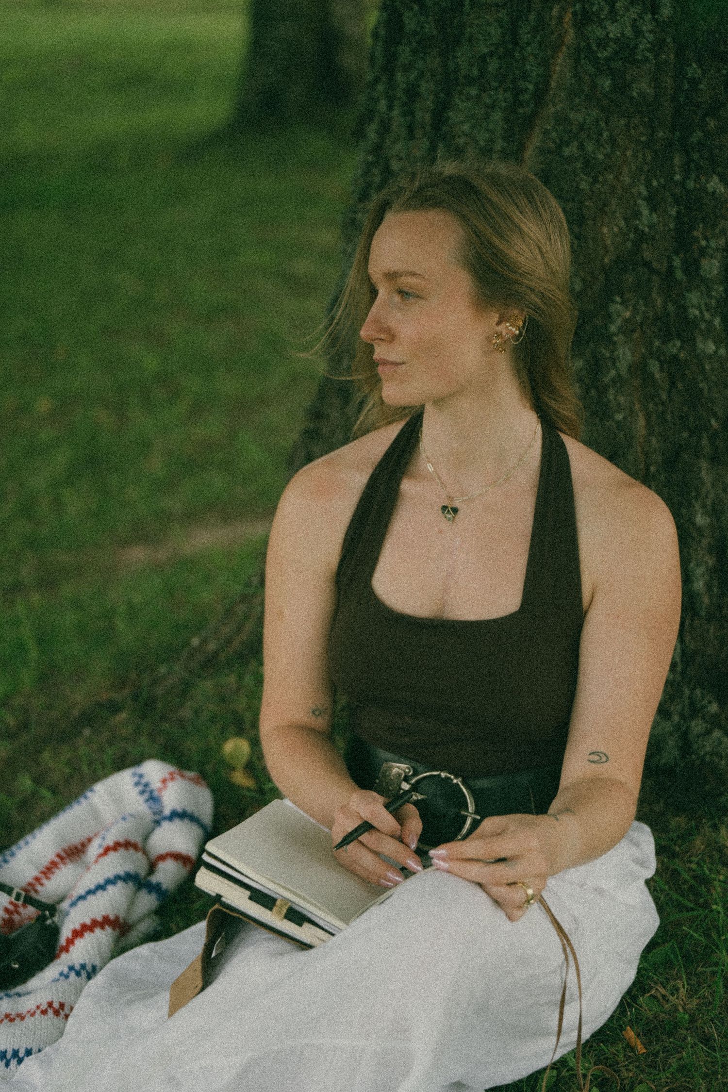Young woman in black halter top sits against tree in park, writing in notebook on sunny day.