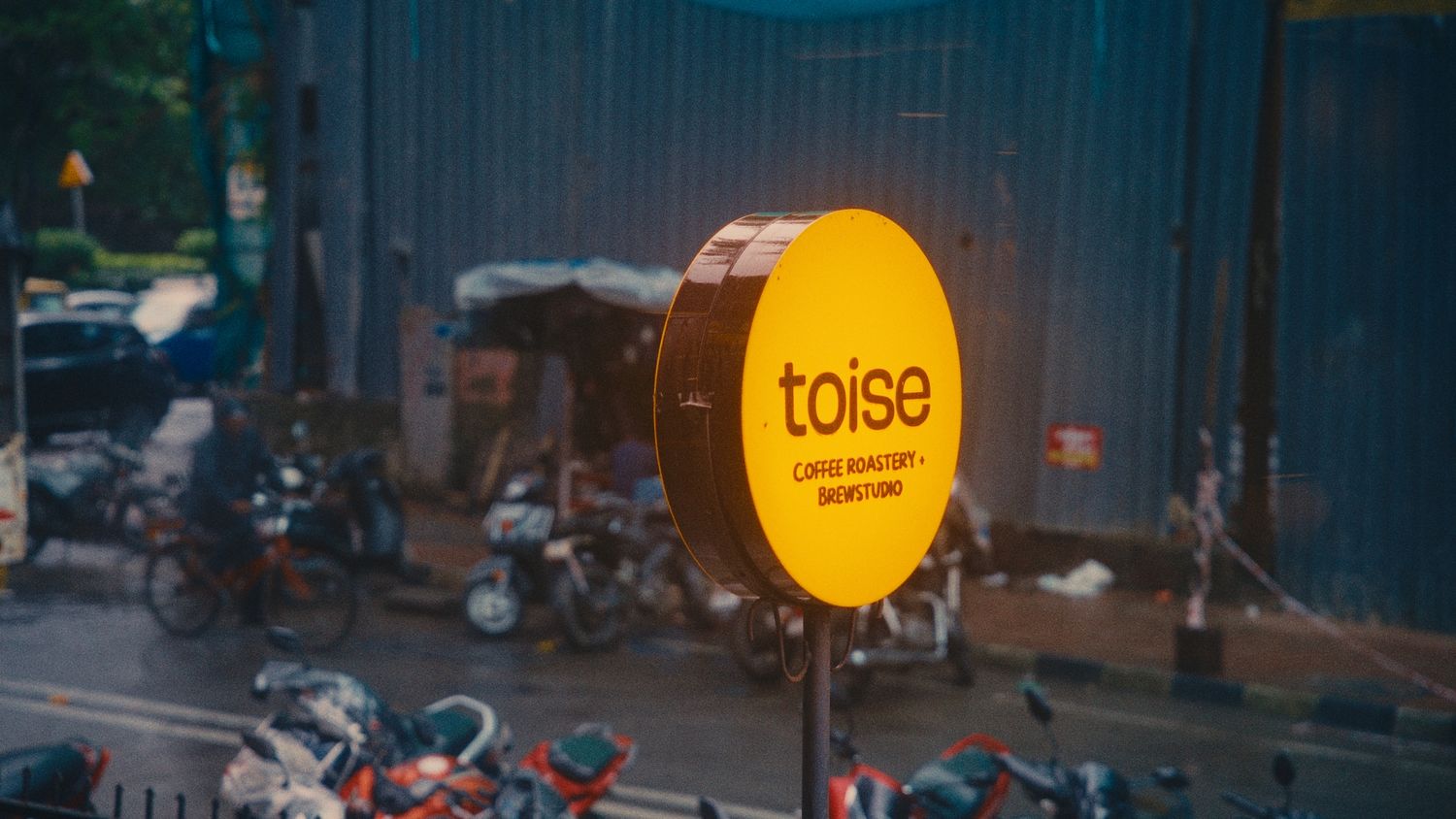 Yellow circular Toise sign glowing on a rainy urban street with motorcycles and blurred city background.