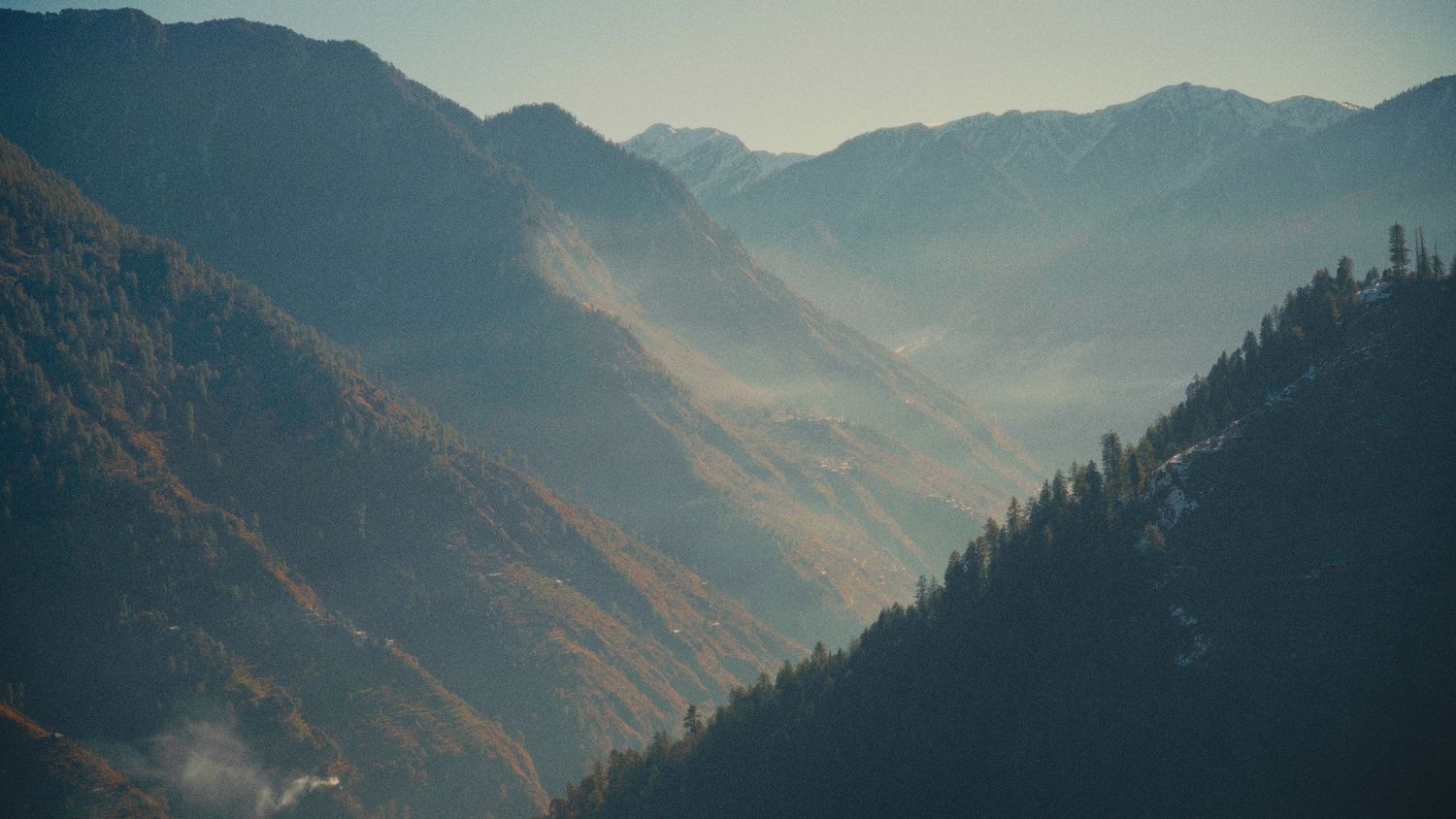 Misty mountain valley with layered peaks, autumn foliage, and evergreen forest in soft hazy light.