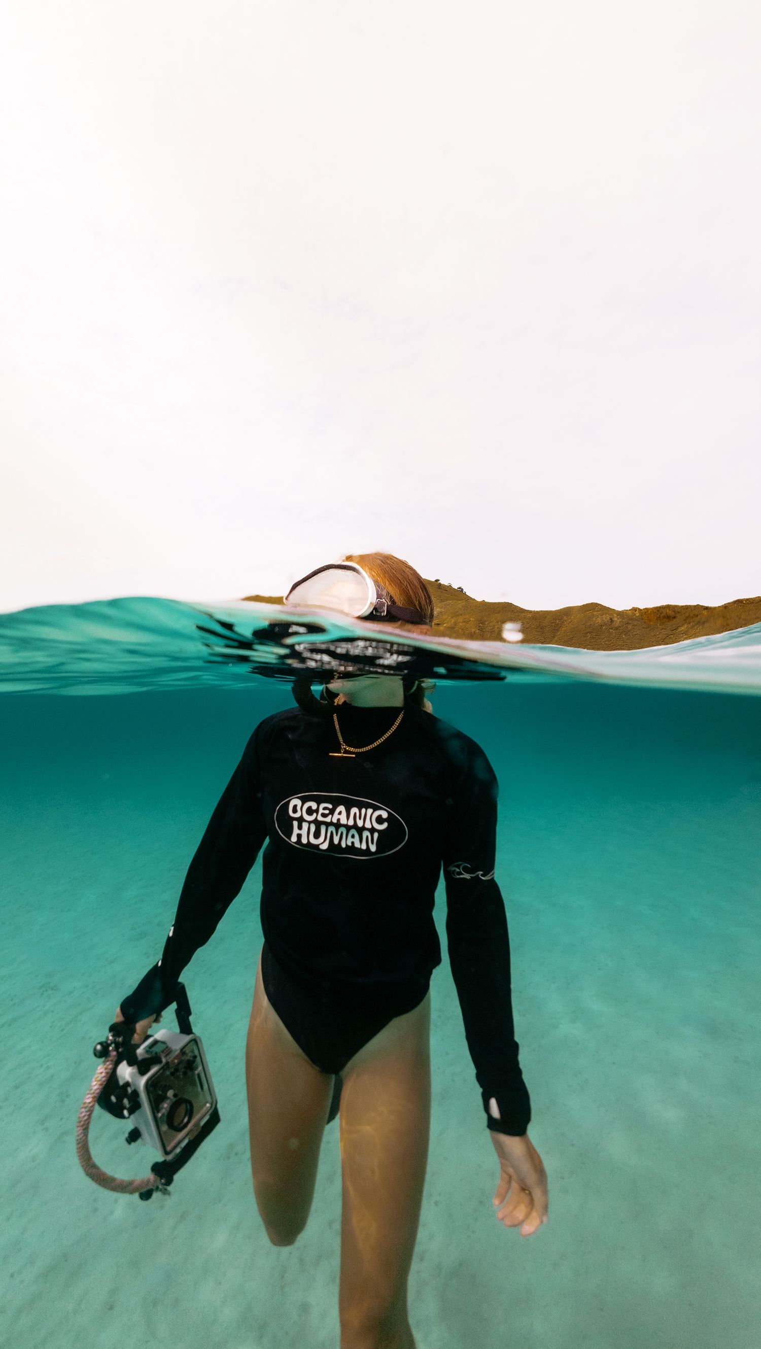Split underwater shot of a swimmer in black wetsuit holding camera equipment in turquoise waters.