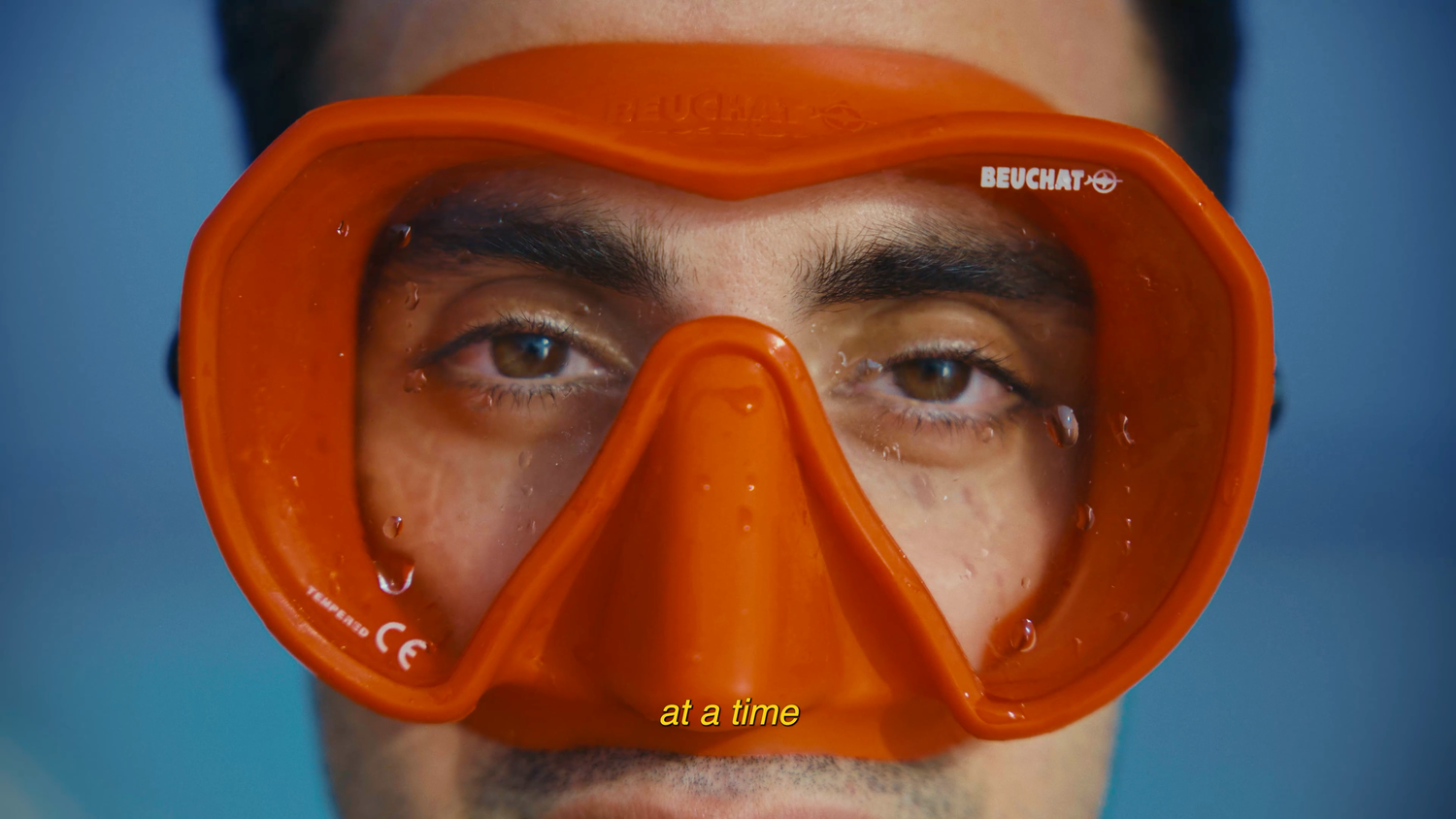 Close-up of a man wearing an orange Beuchat diving mask against a blue background.