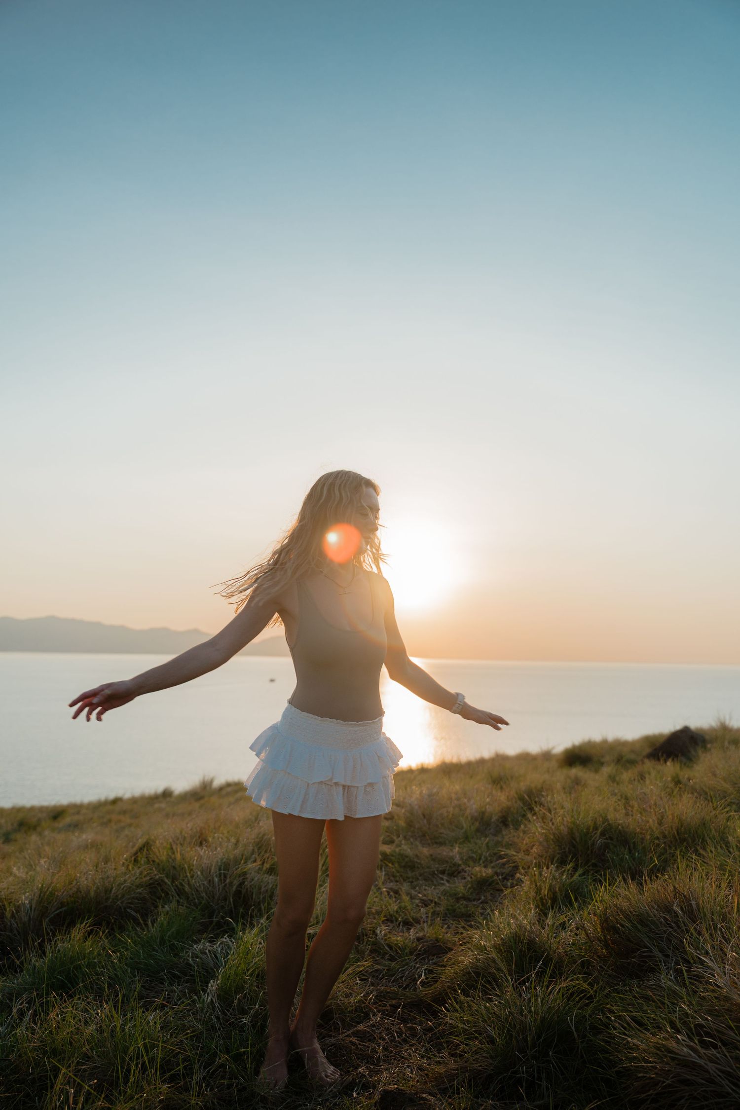 Silhouette of person dancing in golden sunlight on grassy hilltop overlooking ocean.