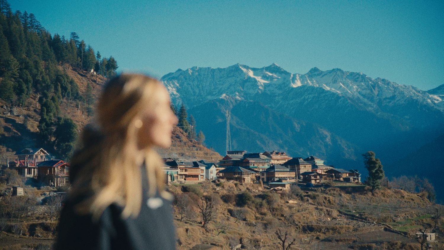 Blonde woman gazing at a scenic Himalayan mountain village with snow-capped peaks under a clear blue sky.