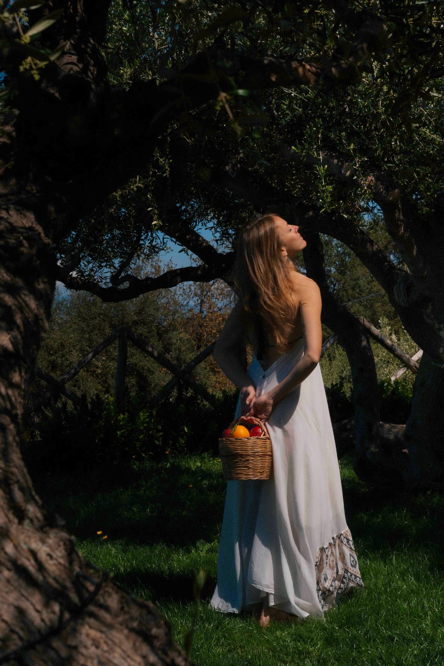 Woman in white dress holding a wicker basket of fruit stands under a large tree in a sunlit garden.