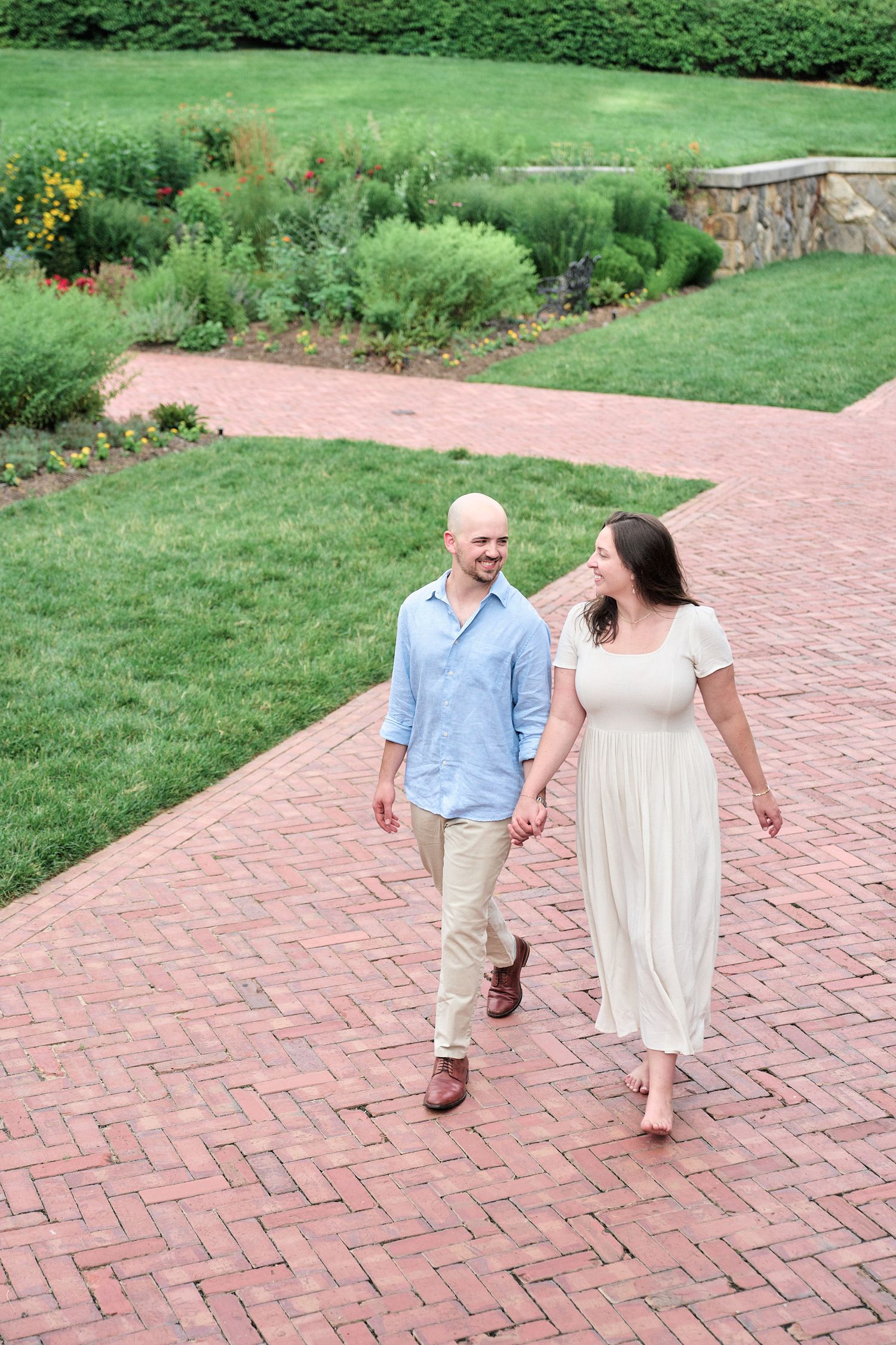 A couple takes a romantic stroll along a winding brick path surrounded by gardens.