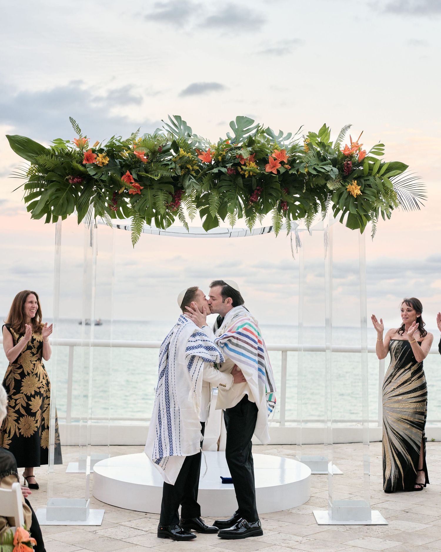 Tropical floral arch with monstera leaves and orange blooms adorns an oceanfront wedding ceremony at sunset.