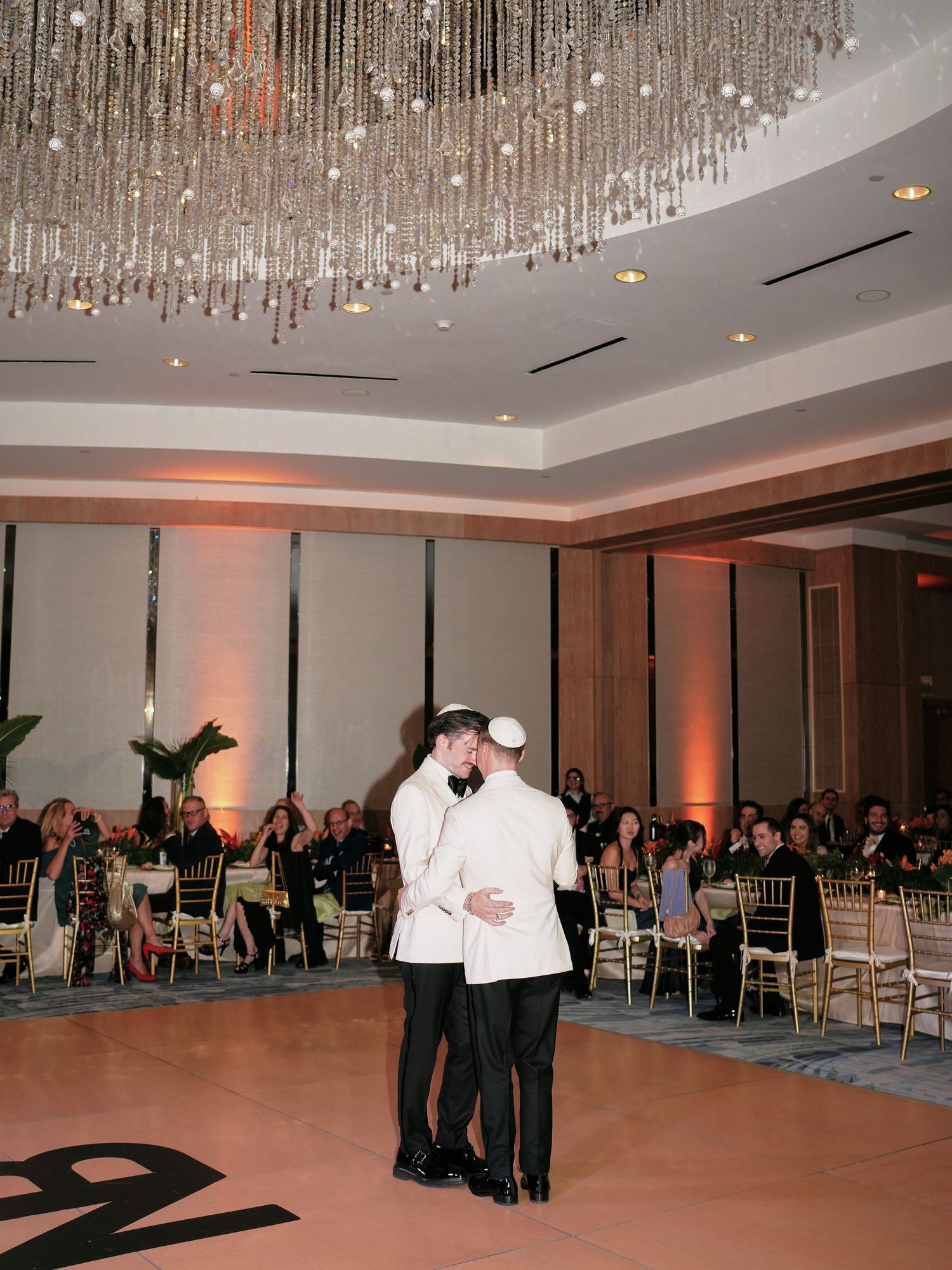 Two people share a romantic first dance beneath a crystal chandelier at an elegant wedding reception ballroom.
