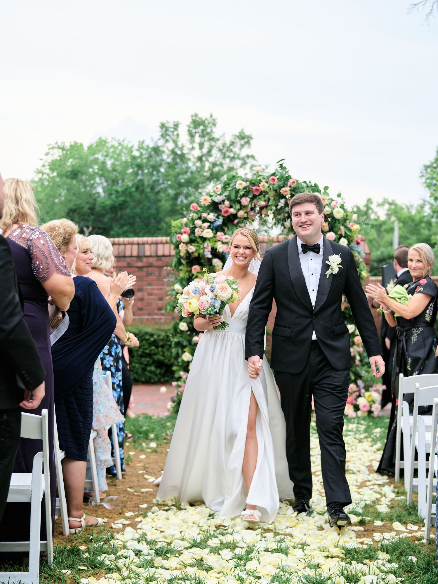 Newlyweds walk down outdoor aisle scattered with rose petals as guests celebrate at garden wedding ceremony.