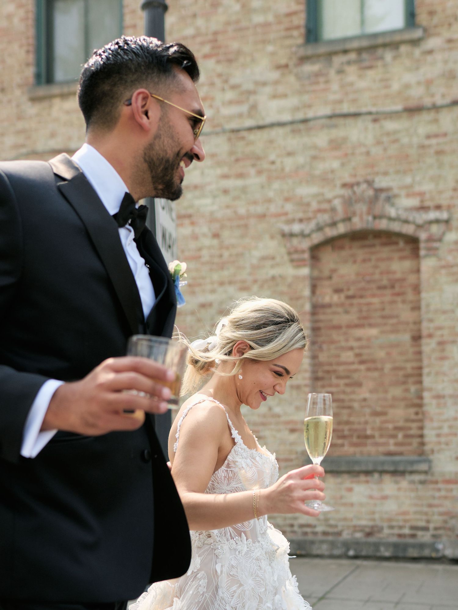 Newlywed couple laughing while holding champagne glasses against a brick building backdrop at their wedding celebration.