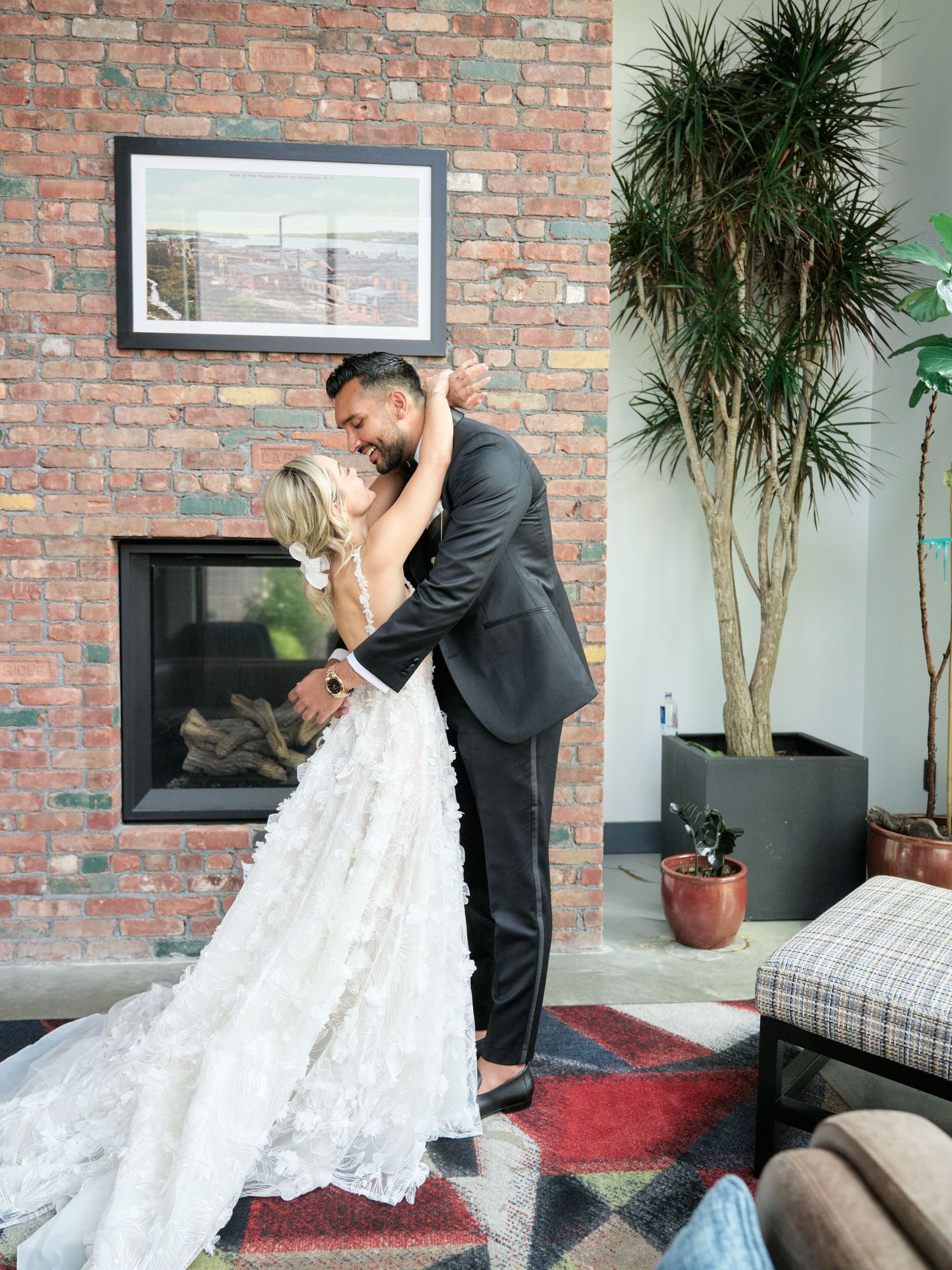 A romantic wedding couple shares a tender moment in front of a brick fireplace with a tall palm plant nearby.