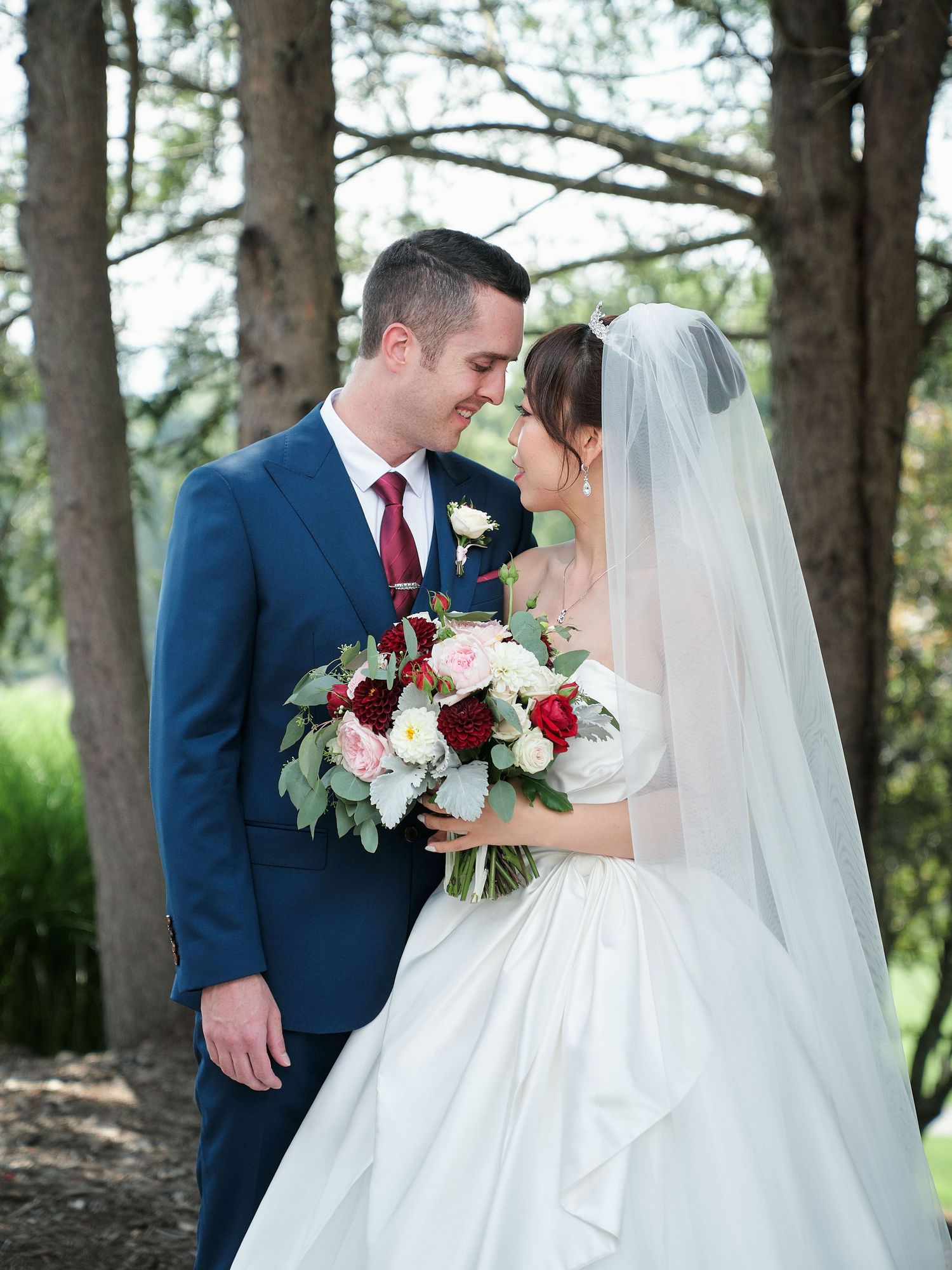 Newlyweds share an intimate moment outdoors with a stunning burgundy and white wedding bouquet.