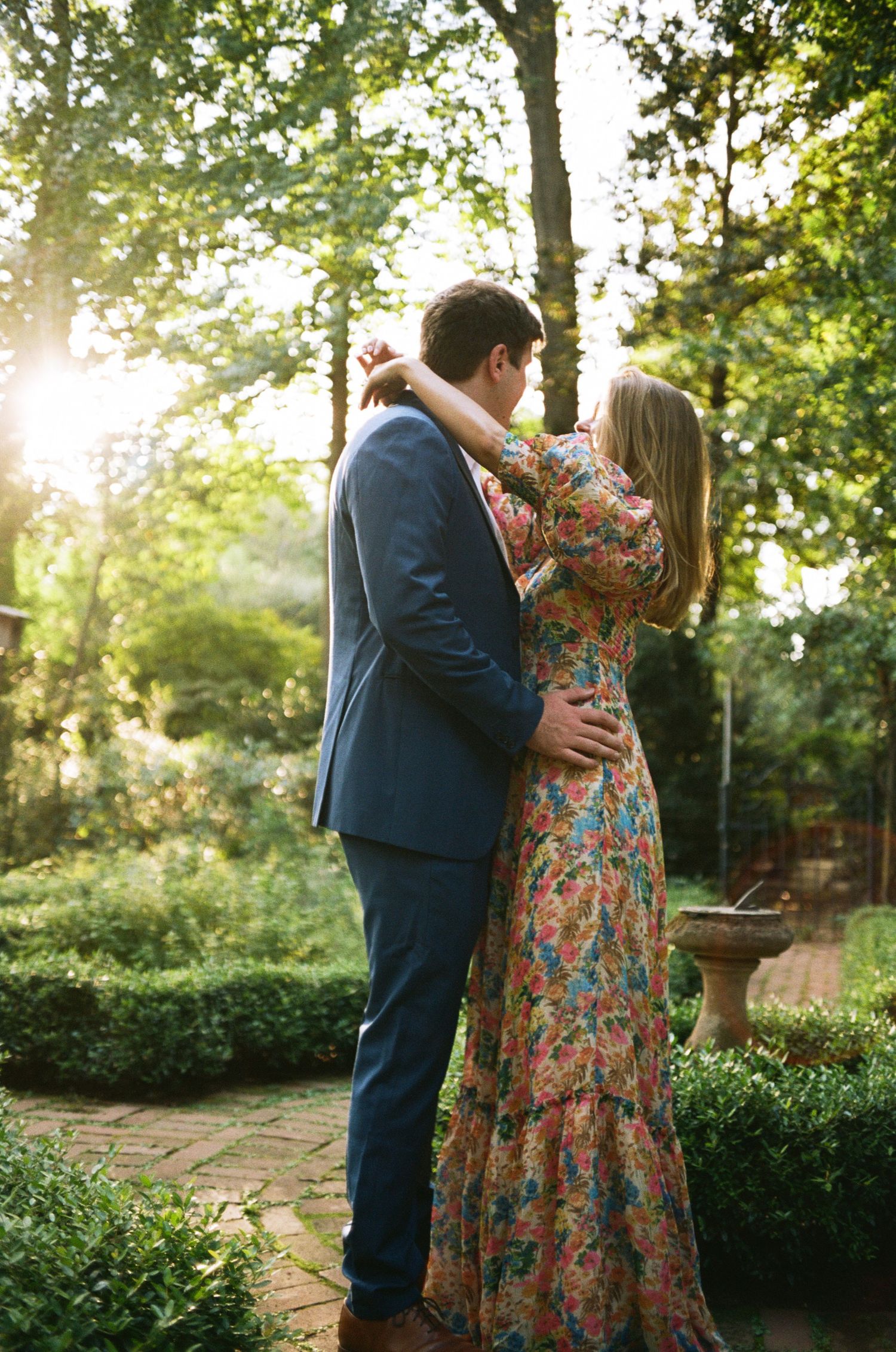 A romantic couple embraces during sunset in a garden setting with golden light filtering through trees.