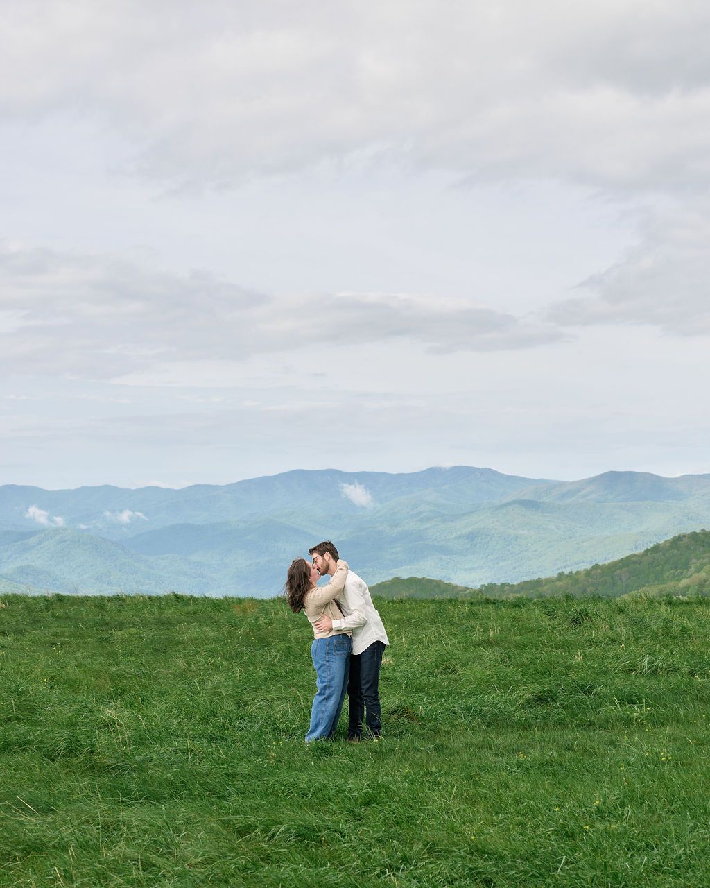 A couple embraces on a grassy hilltop overlooking blue mountain ranges under cloudy skies.
