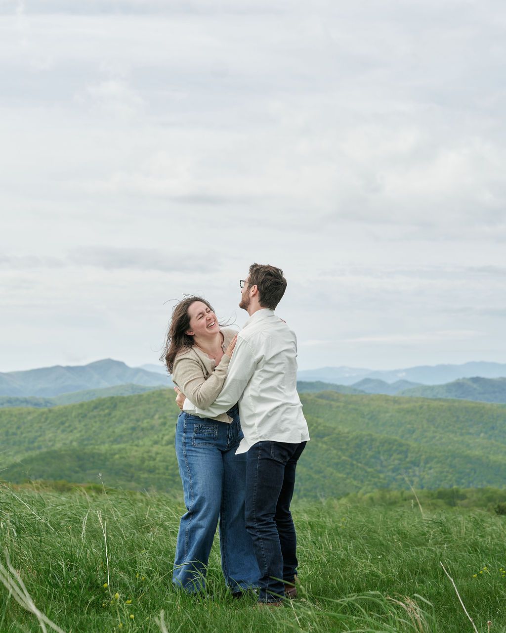 A couple embraces joyfully on a hilltop meadow overlooking rolling green mountains under a cloudy sky.