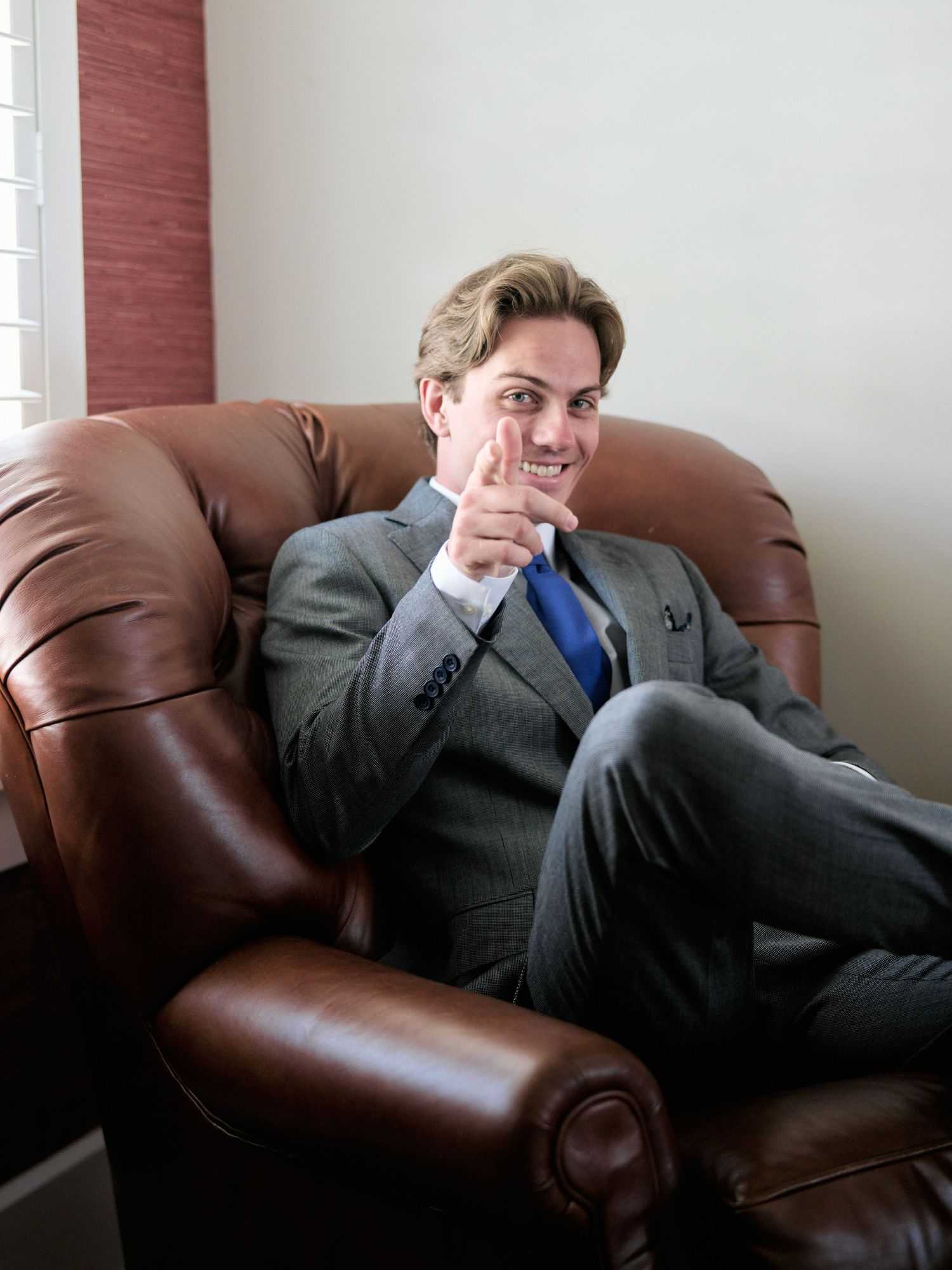 A person in a gray suit and blue tie gives a thumbs up while relaxing on a brown leather armchair.