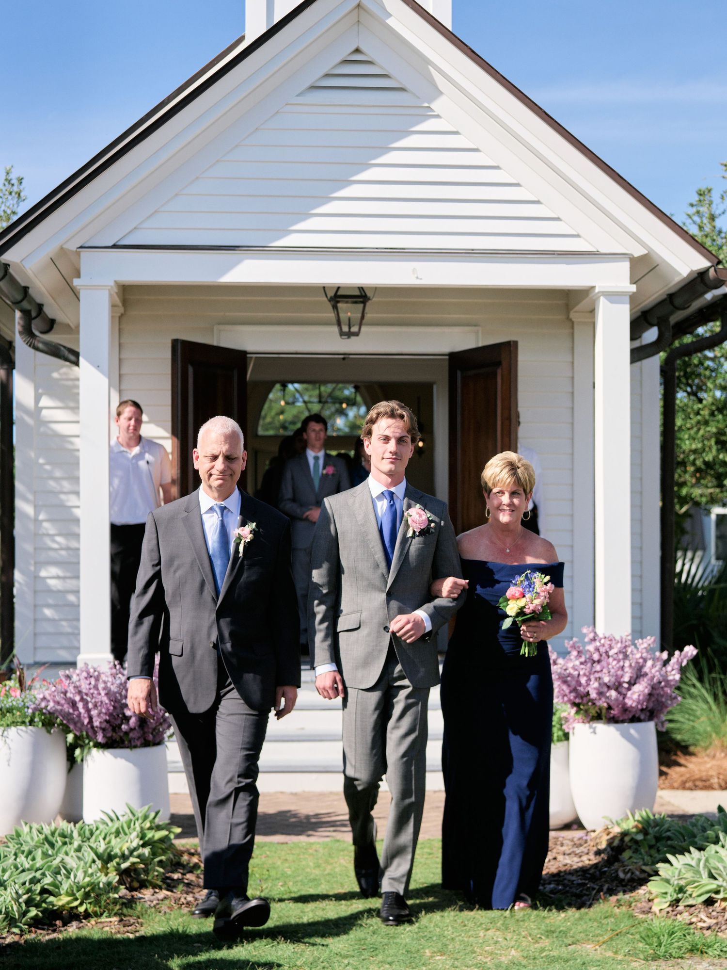 Wedding party exits small white chapel with potted purple flowers flanking the entrance on a sunny day.