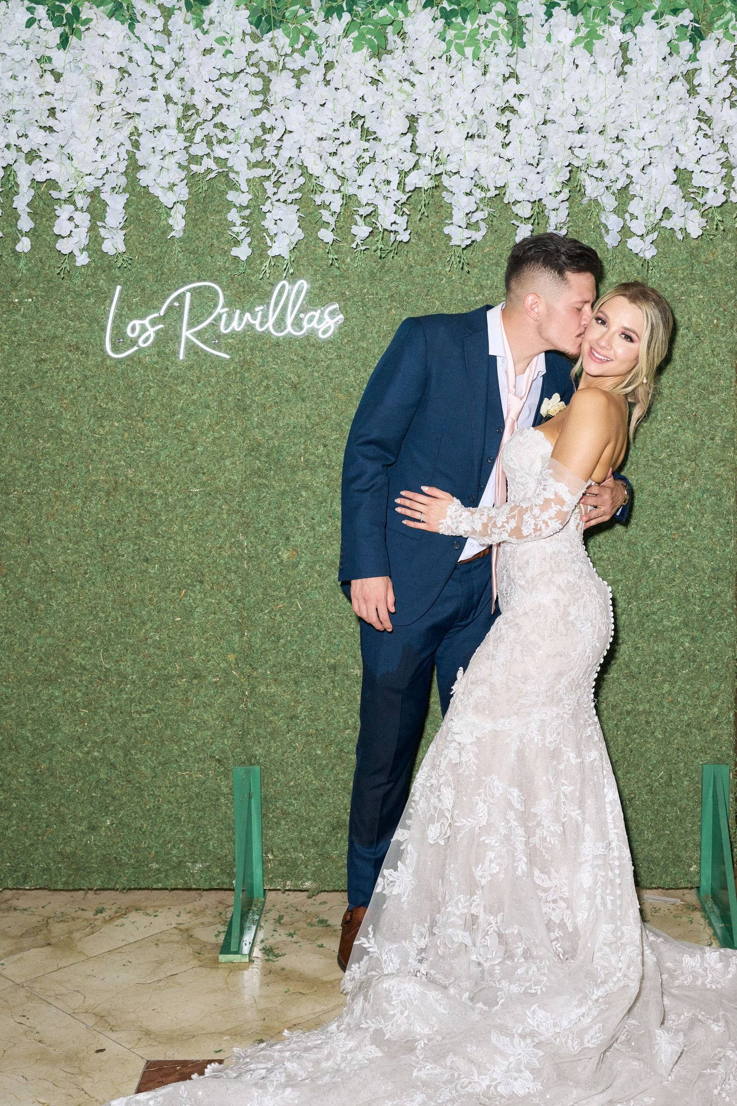 Wedding couple embraces in front of green wall decorated with cascading white flowers and neon Los Ruellos sign.