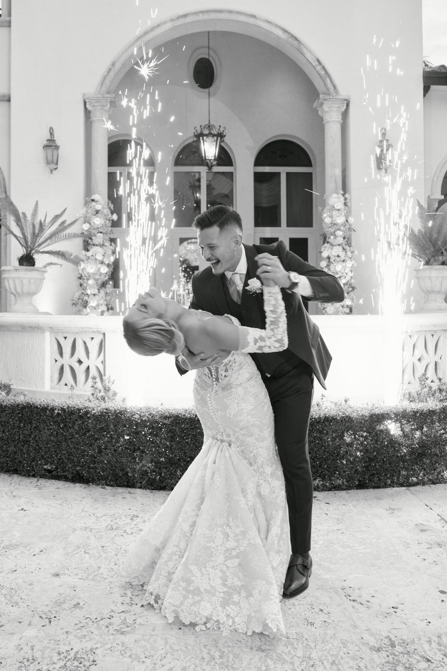 Black and white photo of a romantic dip pose in front of an elegant archway with white floral decor.