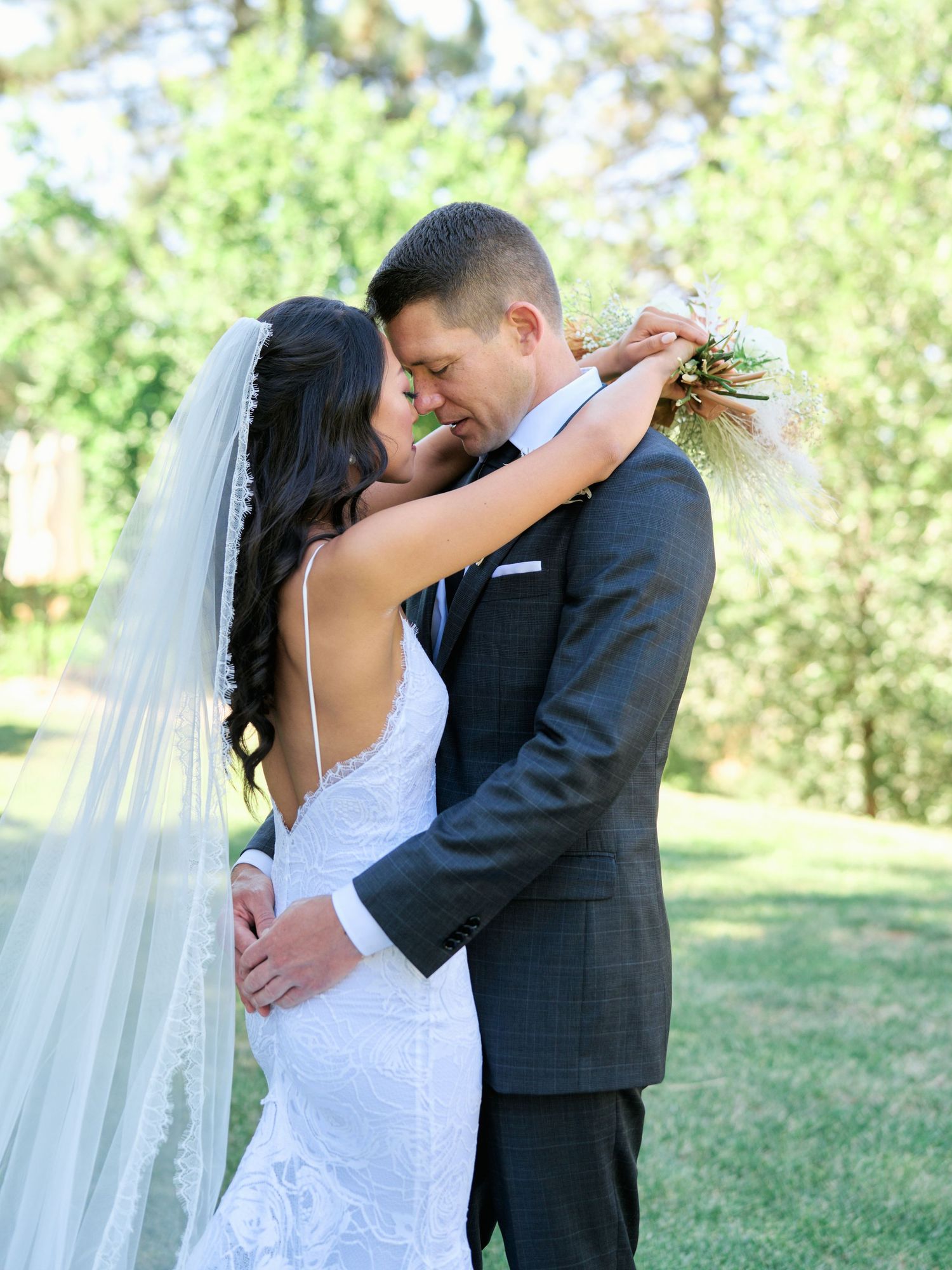 A romantic wedding moment captured outdoors as a couple in formal attire embraces beneath sunlit trees.