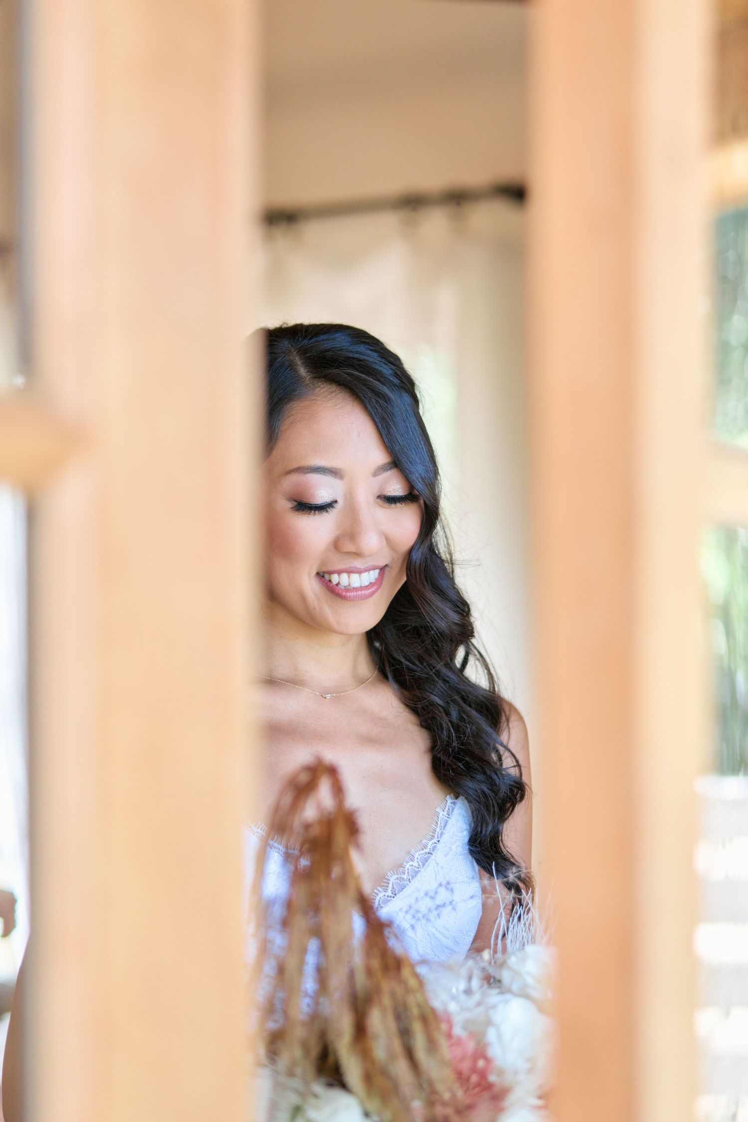 Candid moment of a bride in a white dress smiling through a window frame.