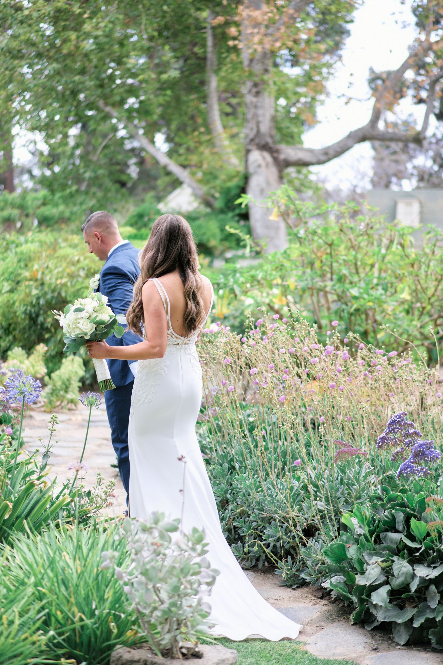 Wedding couple walking along garden path surrounded by blooming flowers and greenery.