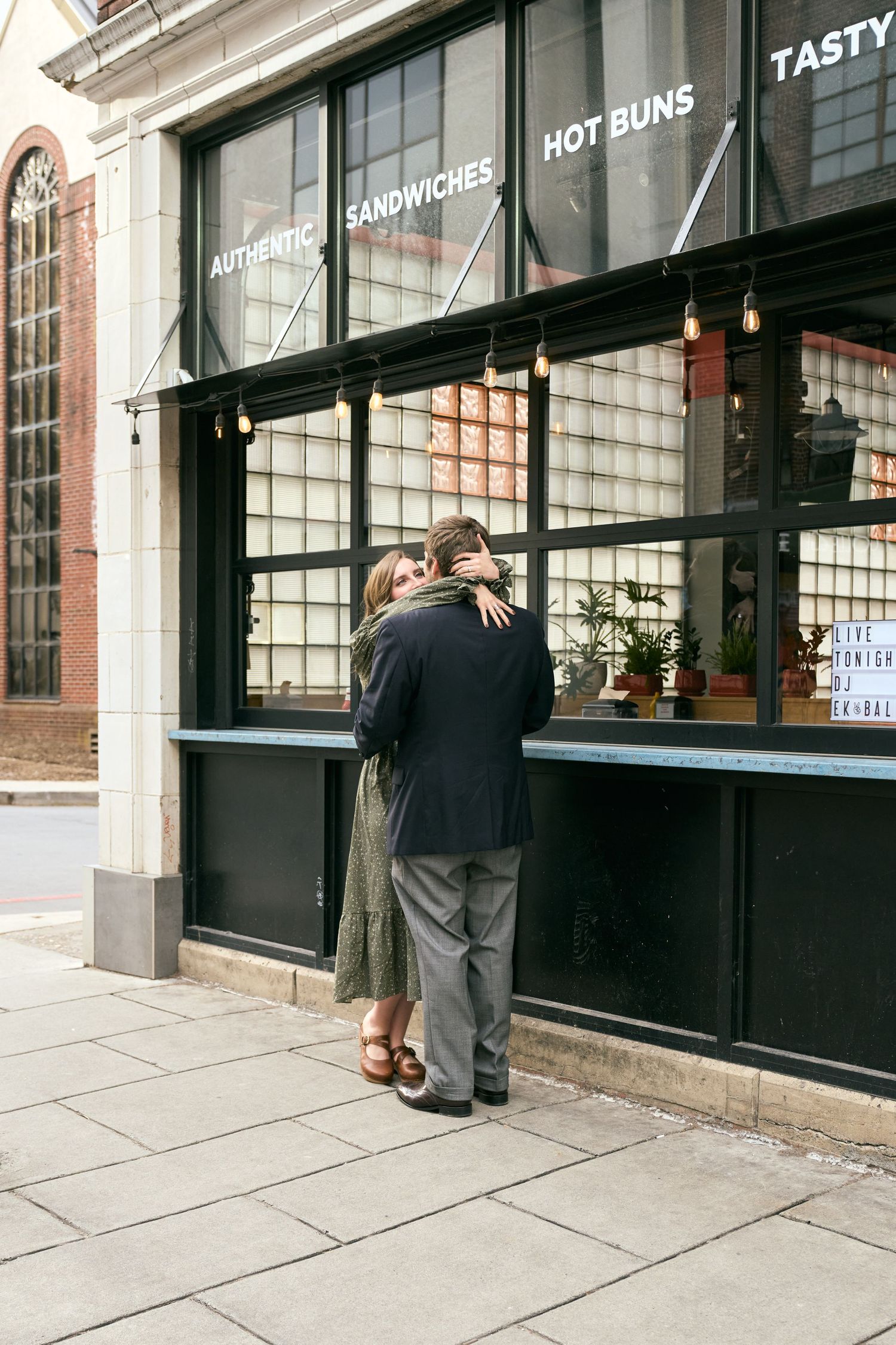 A couple embraces outside a storefront with 'Hot Buns' signage.
