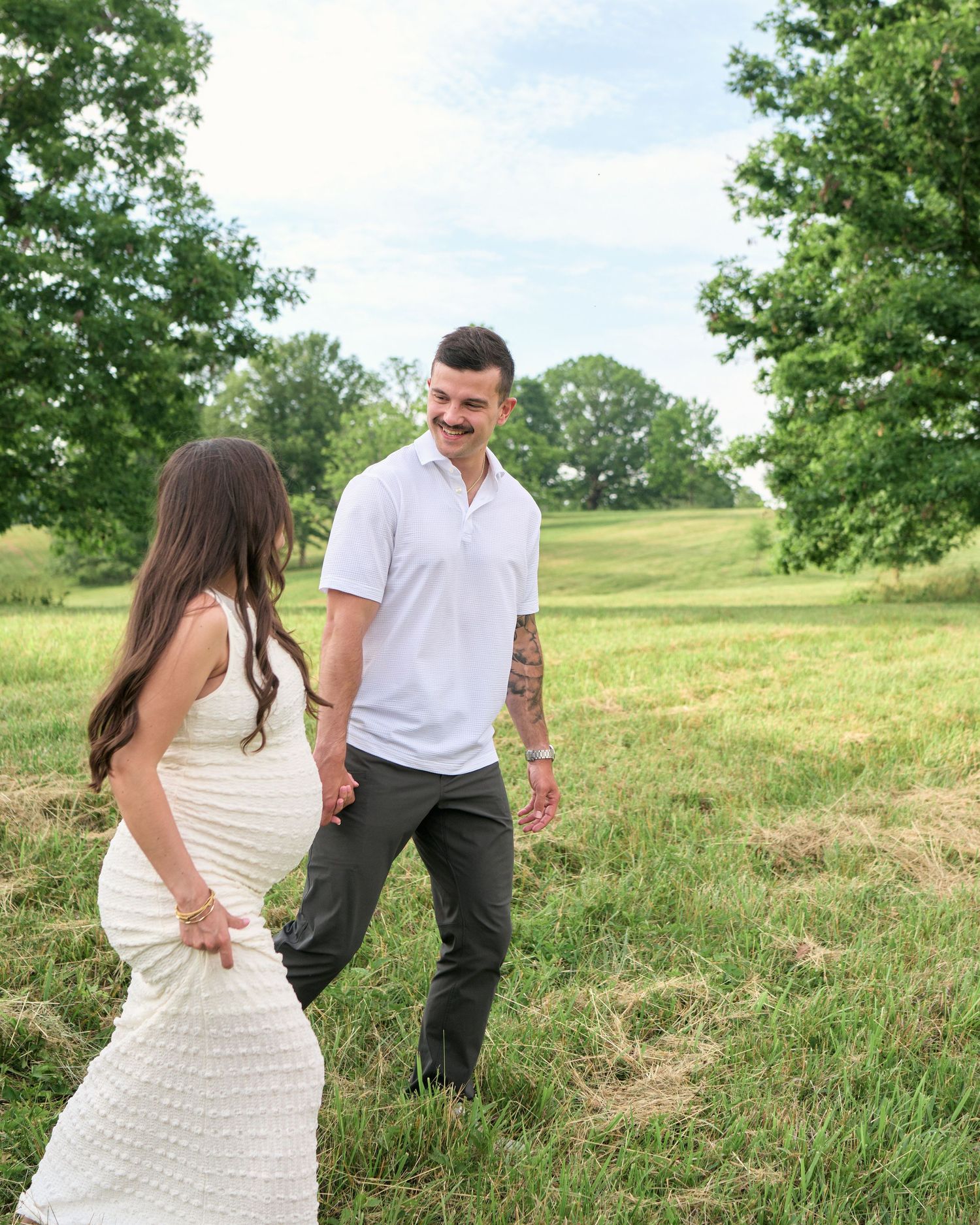 Couple holding hands in a green field, woman in white dress, man in white polo with tattoos, outdoor maternity photo.