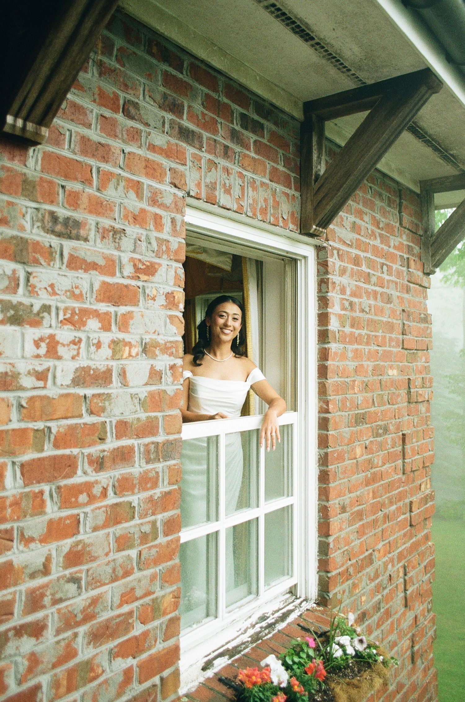 Smiling bride in white dress leaning out of a brick building window with flower box below.