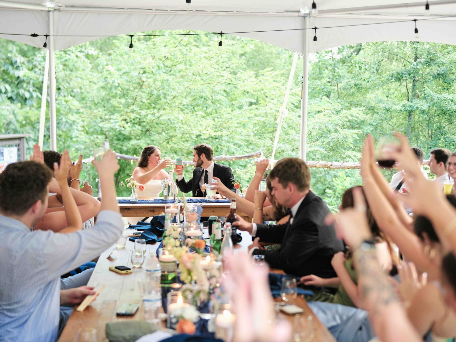 Wedding reception toast under a tent with guests raising glasses toward the bride and groom at a long decorated table.