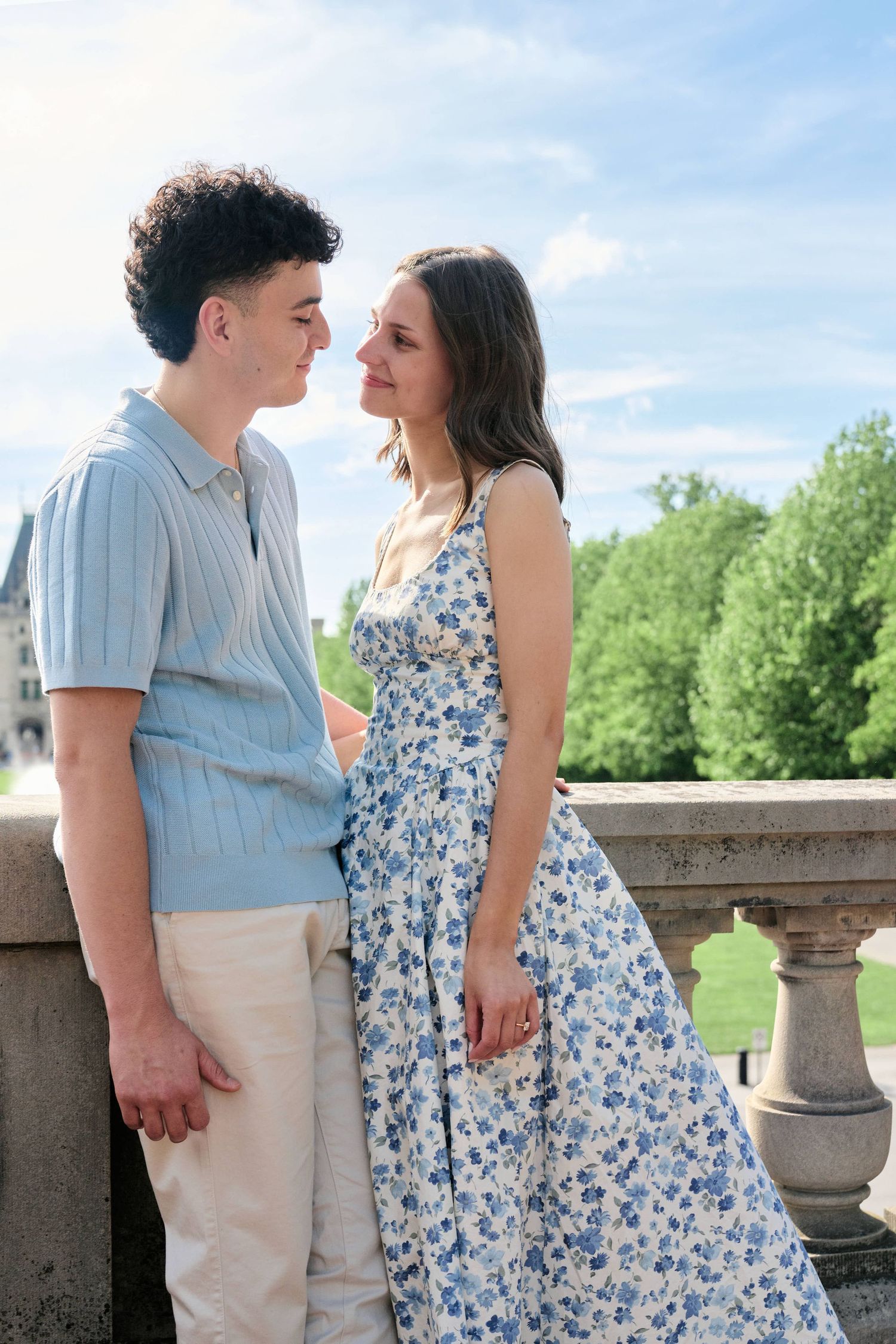 Young couple gazing into each other's eyes on a stone balcony, man in blue polo and woman in floral dress.