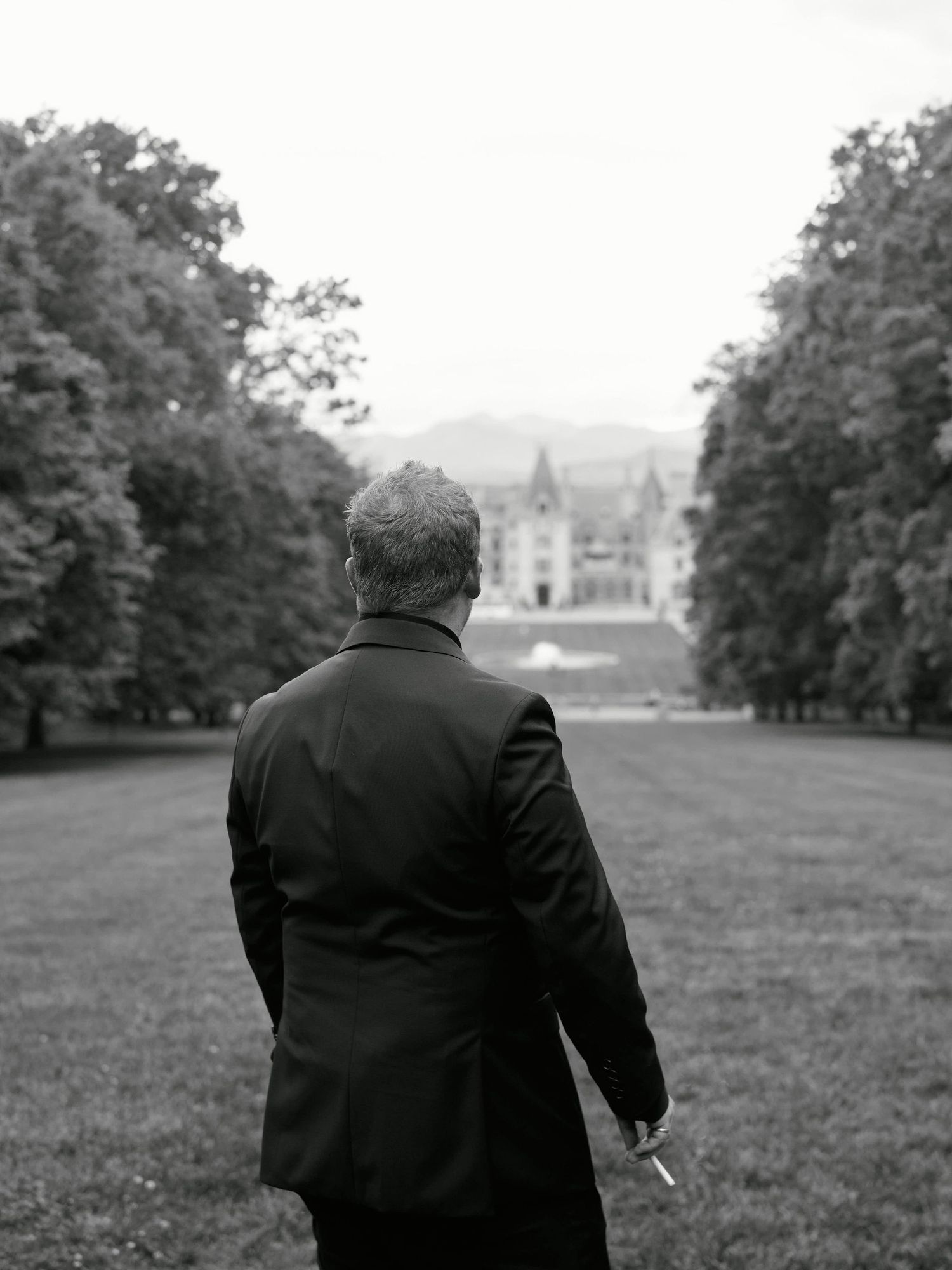 A person in black clothing walks away from the camera across a grassy field with trees in the background.