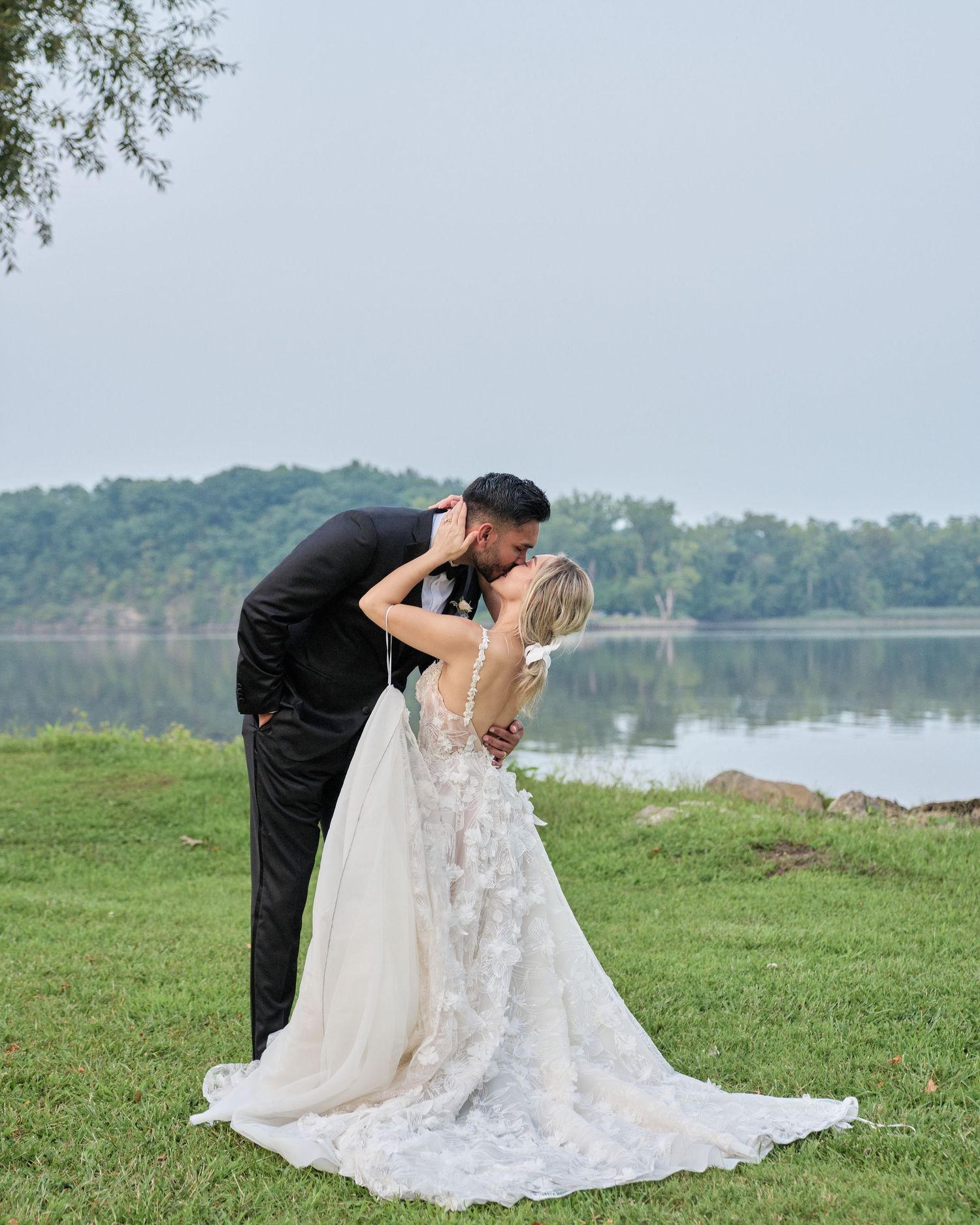 A bride and groom embrace each other outdoors in a grassy field near a body of water.