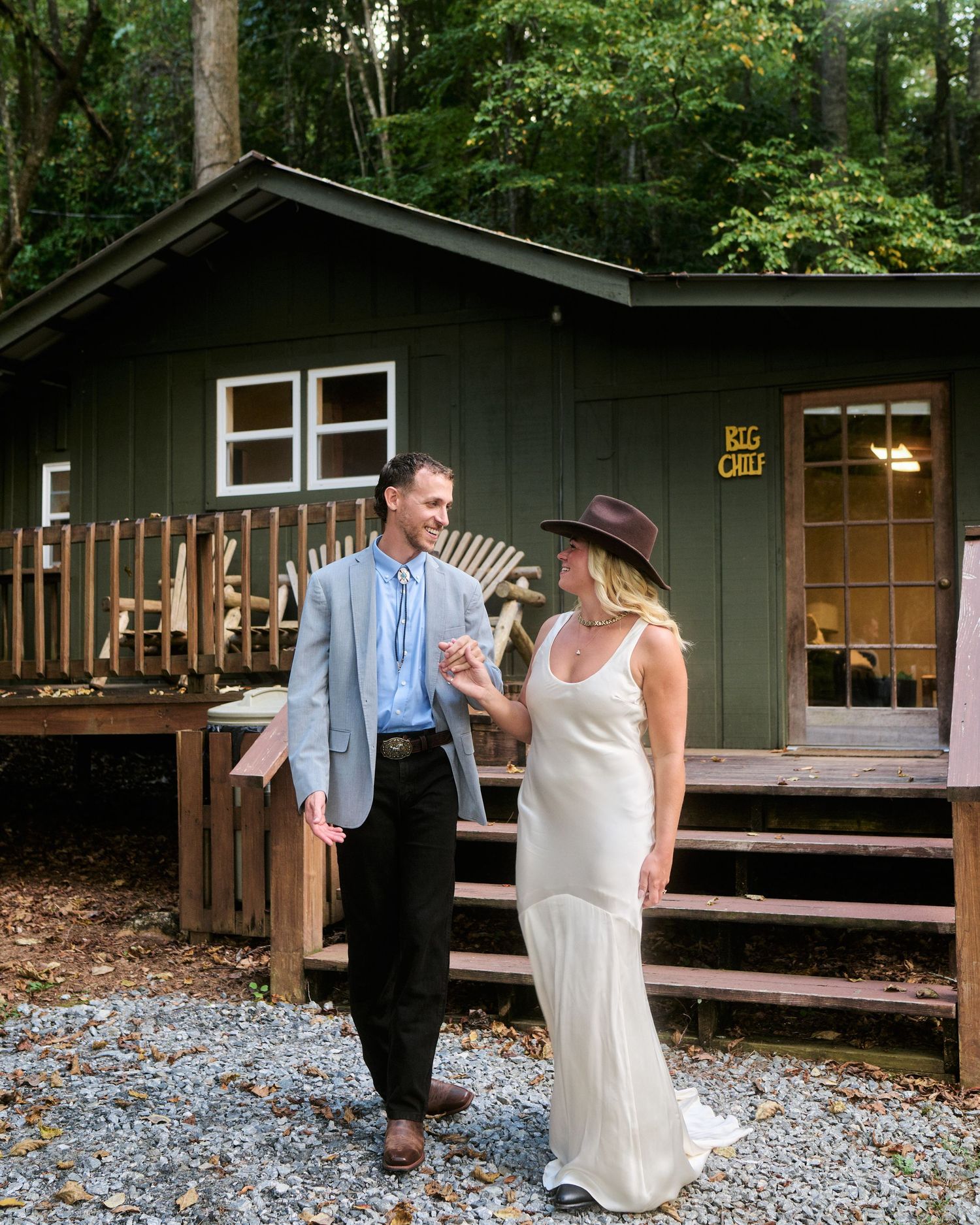 A couple shares a romantic moment outside rustic wooden cabins surrounded by lush forest scenery.