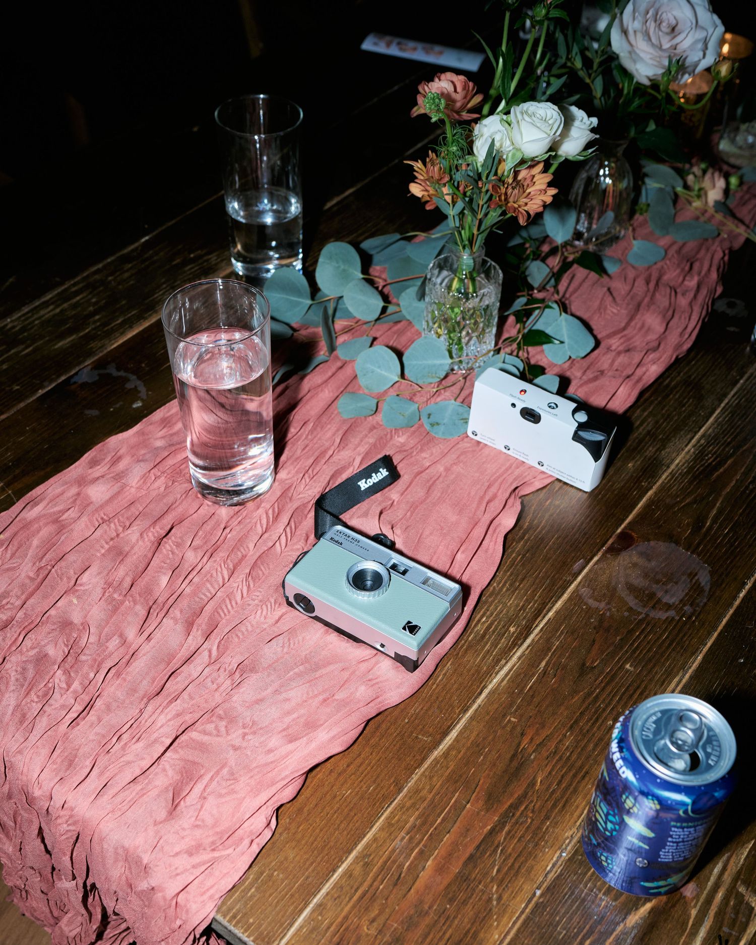 Rustic wedding table with Kodak disposable cameras, pink table runner, floral centerpiece, water glass, and beer can.