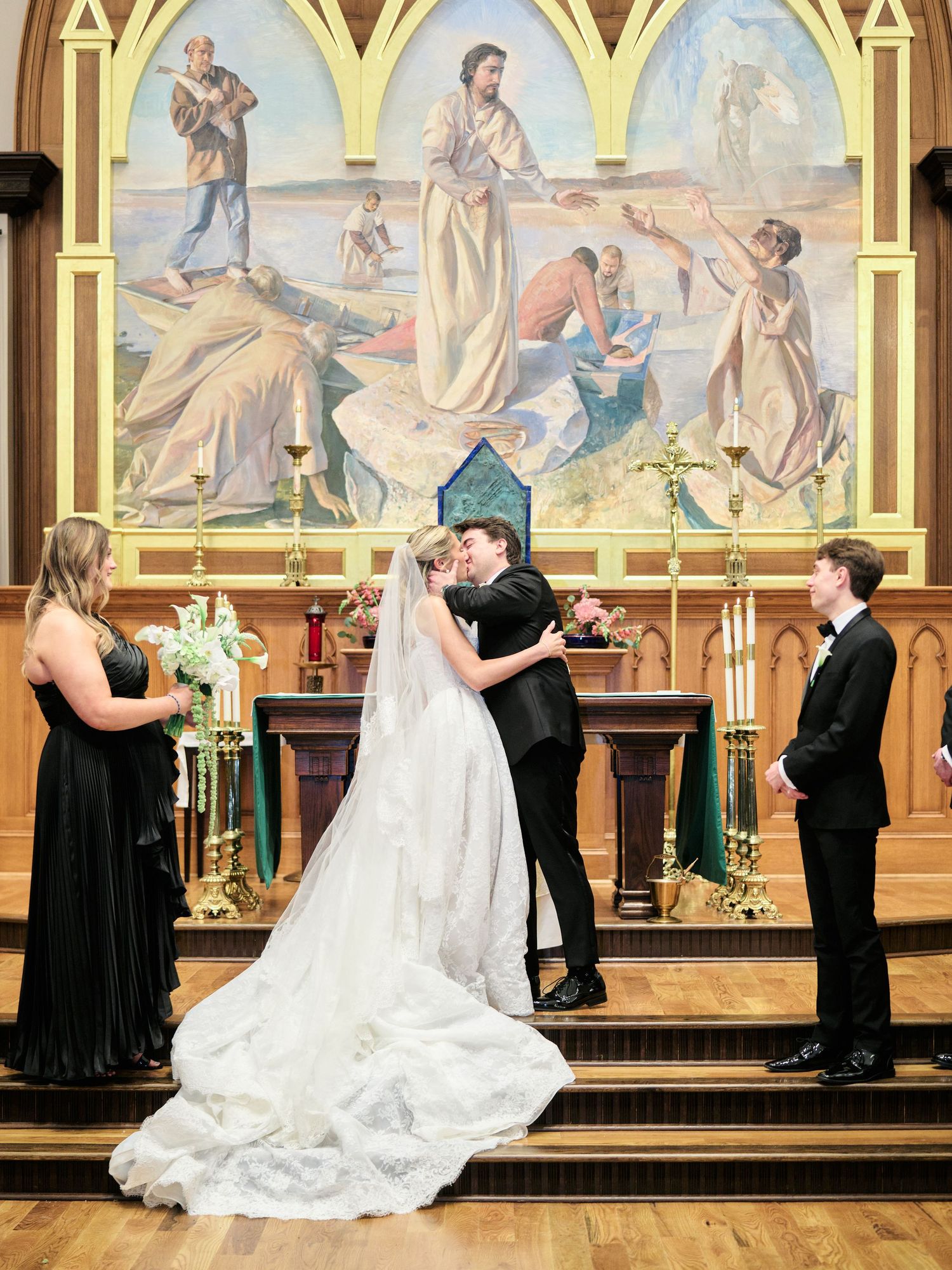 Bride and groom share first kiss at church altar, flanked by wedding party, with religious mural backdrop.