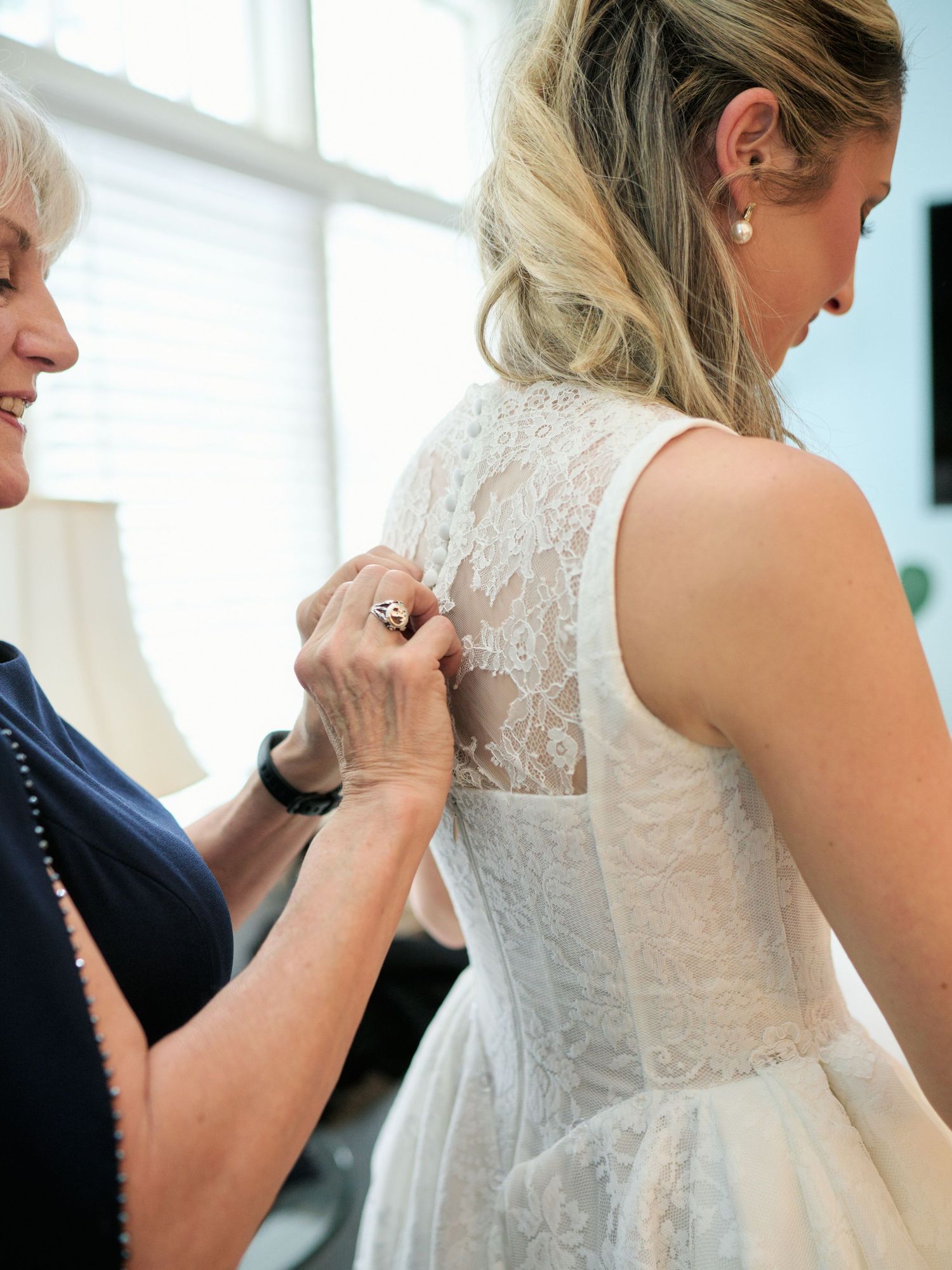 An older woman helps button the back of a brides white lace wedding dress before the ceremony.