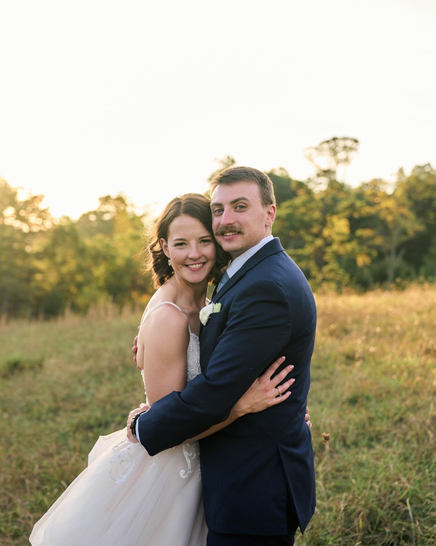 A bride and groom embrace in a golden sunlit field, smiling warmly in wedding attire at an outdoor ceremony.