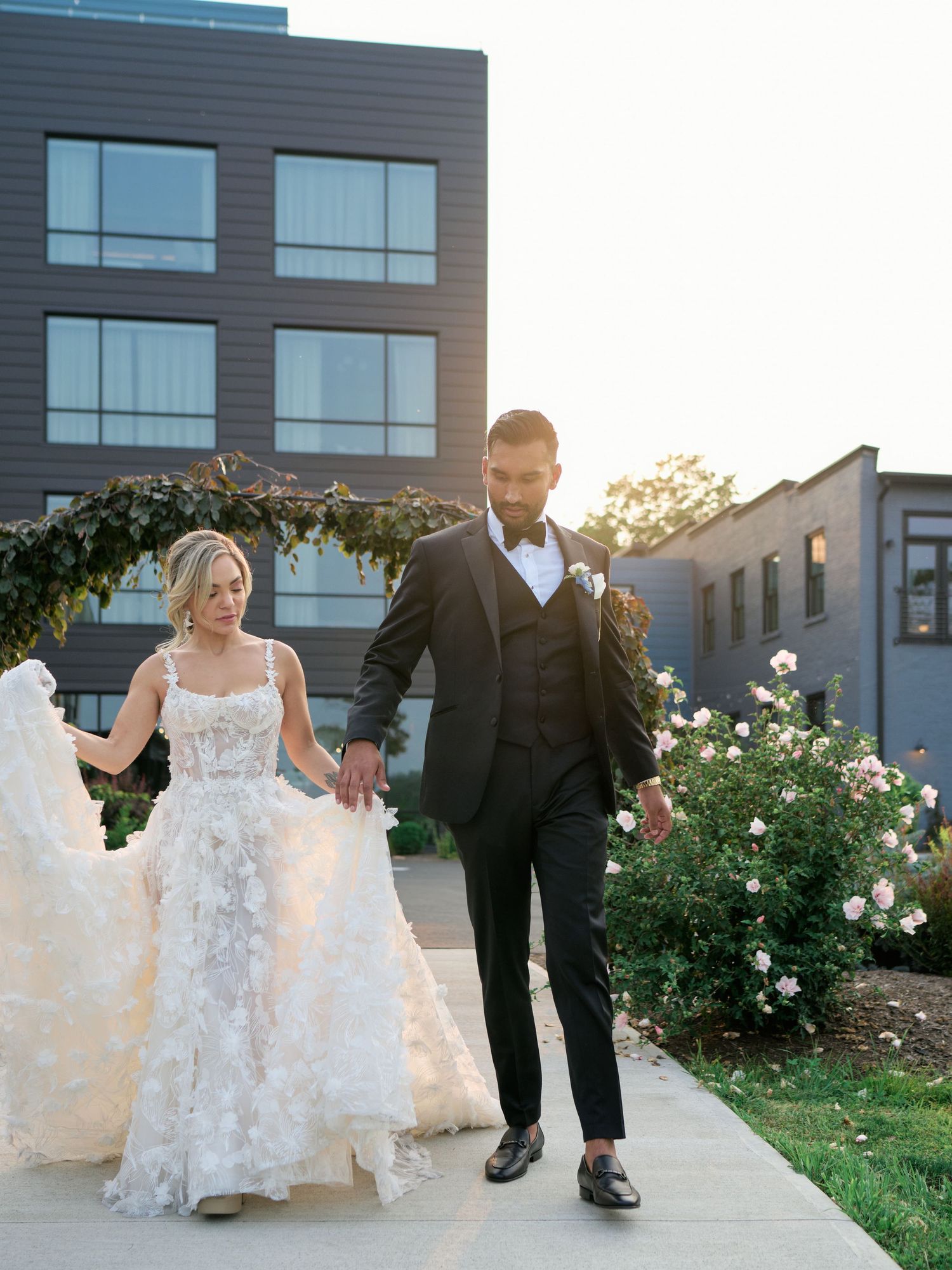 A newlywed couple walks together on a garden path at sunset with white roses and modern buildings in the background.